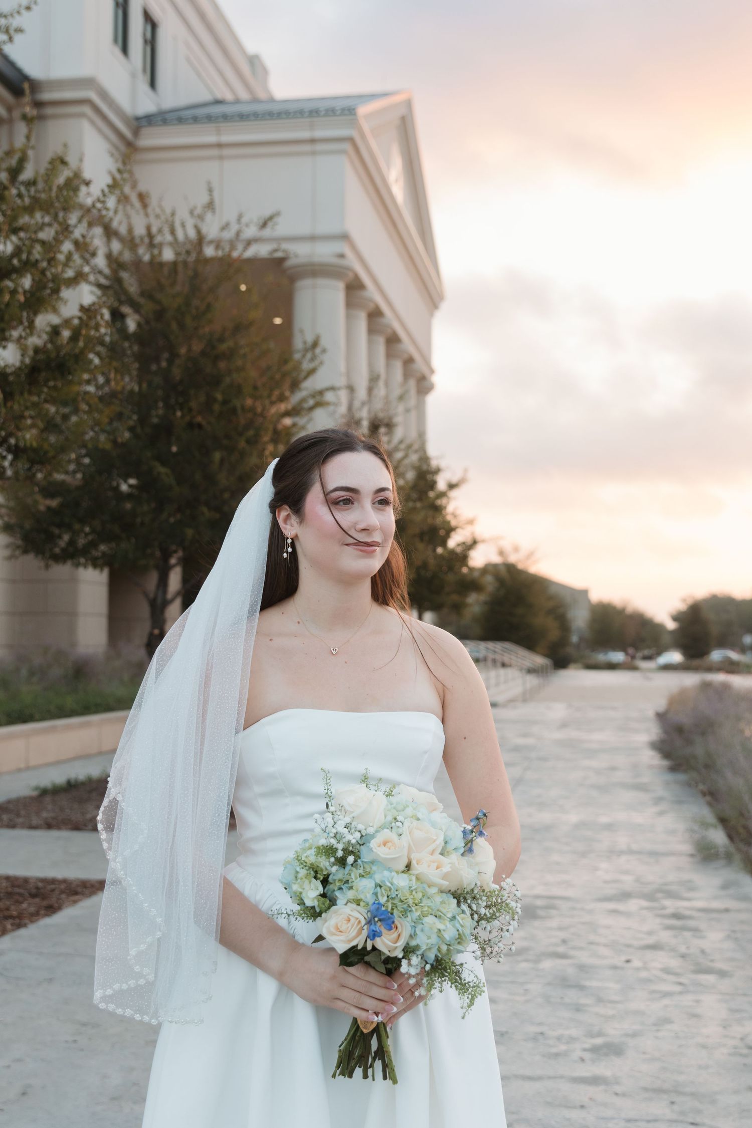 A bride in a white strapless gown holds a delicate bouquet  against a sunset backdrop. Wedding Photographer Fort Worth