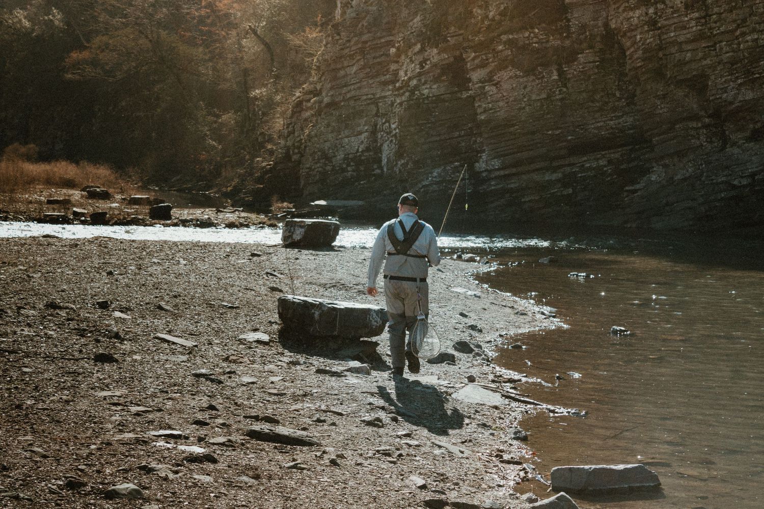 A fisher walks along a rocky riverbank beneath tall cliffs.
