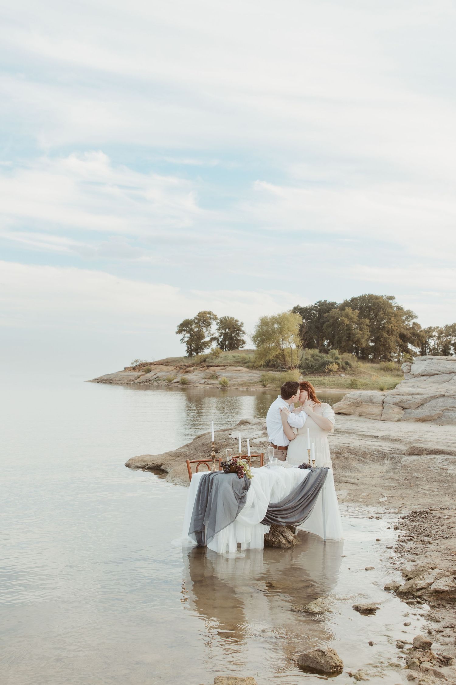 A romantic lakeside picnic setup with white linens and a couple in light clothing on a rocky shore.