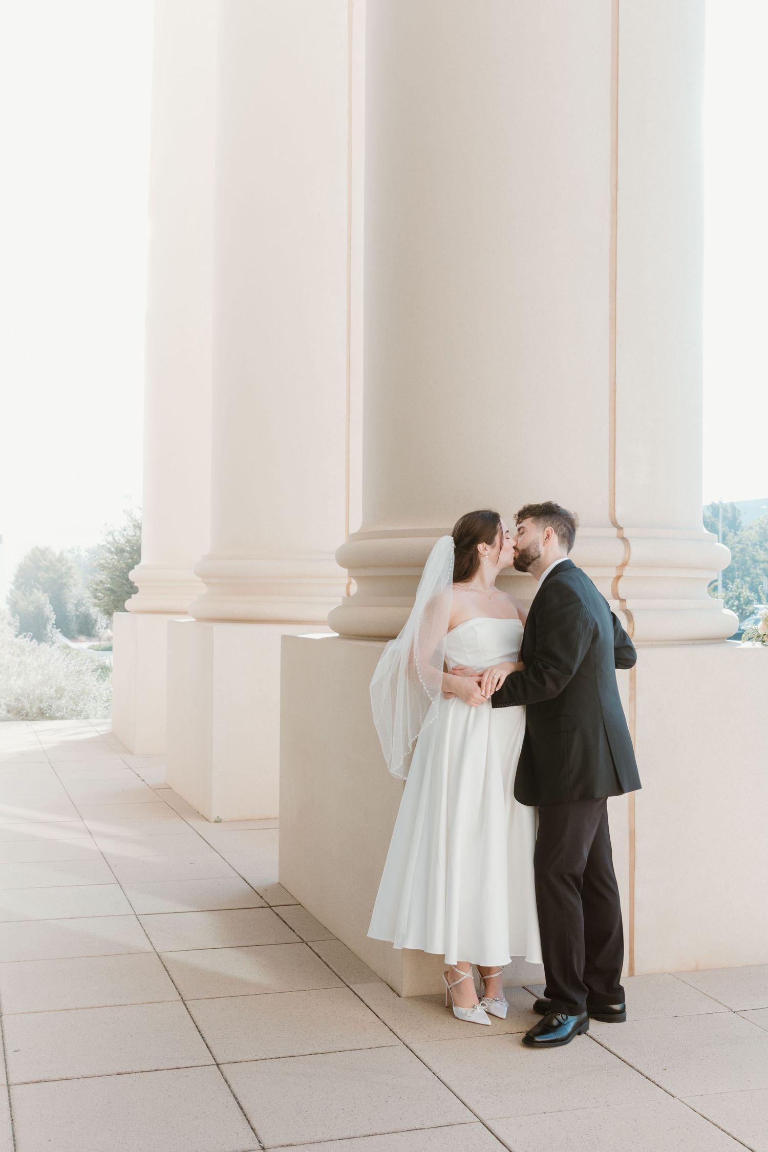 A wedding couple embraces in a bright, minimalist setting with white columns and tiled floor. Wedding Photographer Fort Worth