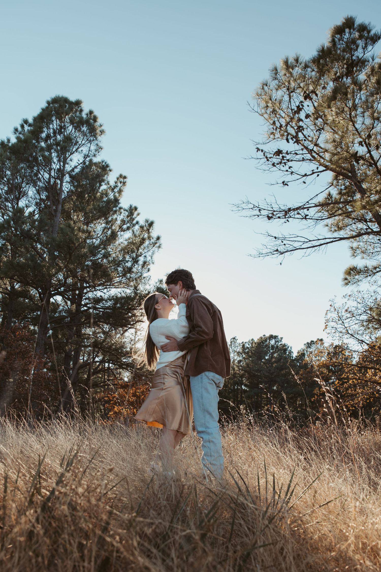 A couple shares romantic moments in a field with pine trees and golden grass in the background. engagement photos DFW