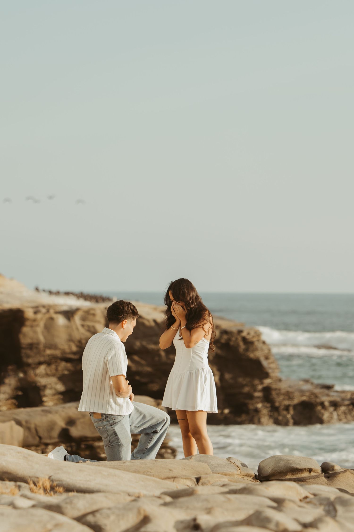 Man proposing on one knee to woman in white dress on rocky ocean shore at sunset.