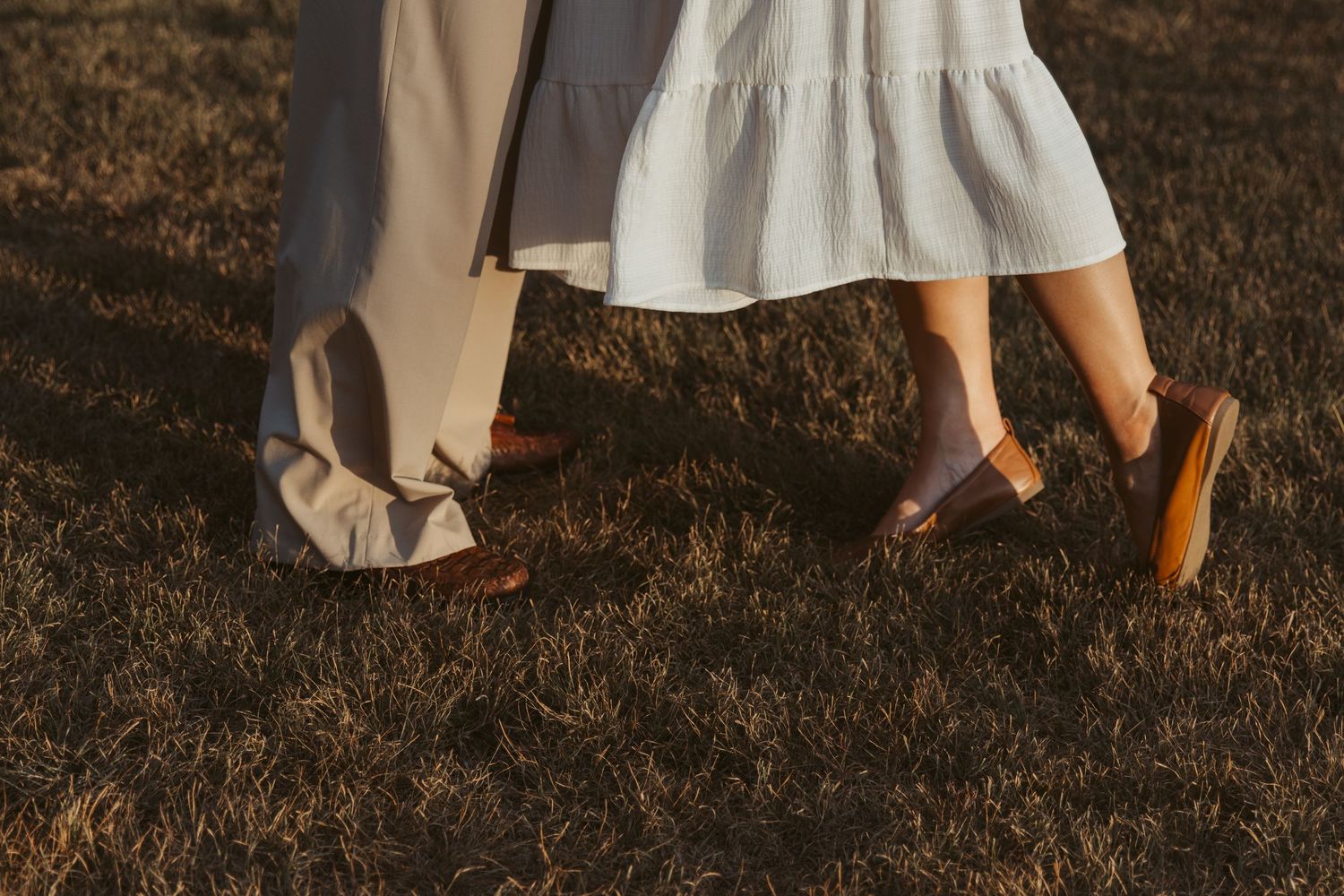 Close-up of two pairs of feet standing on dry grass with white dress hem and casual shoes visible.