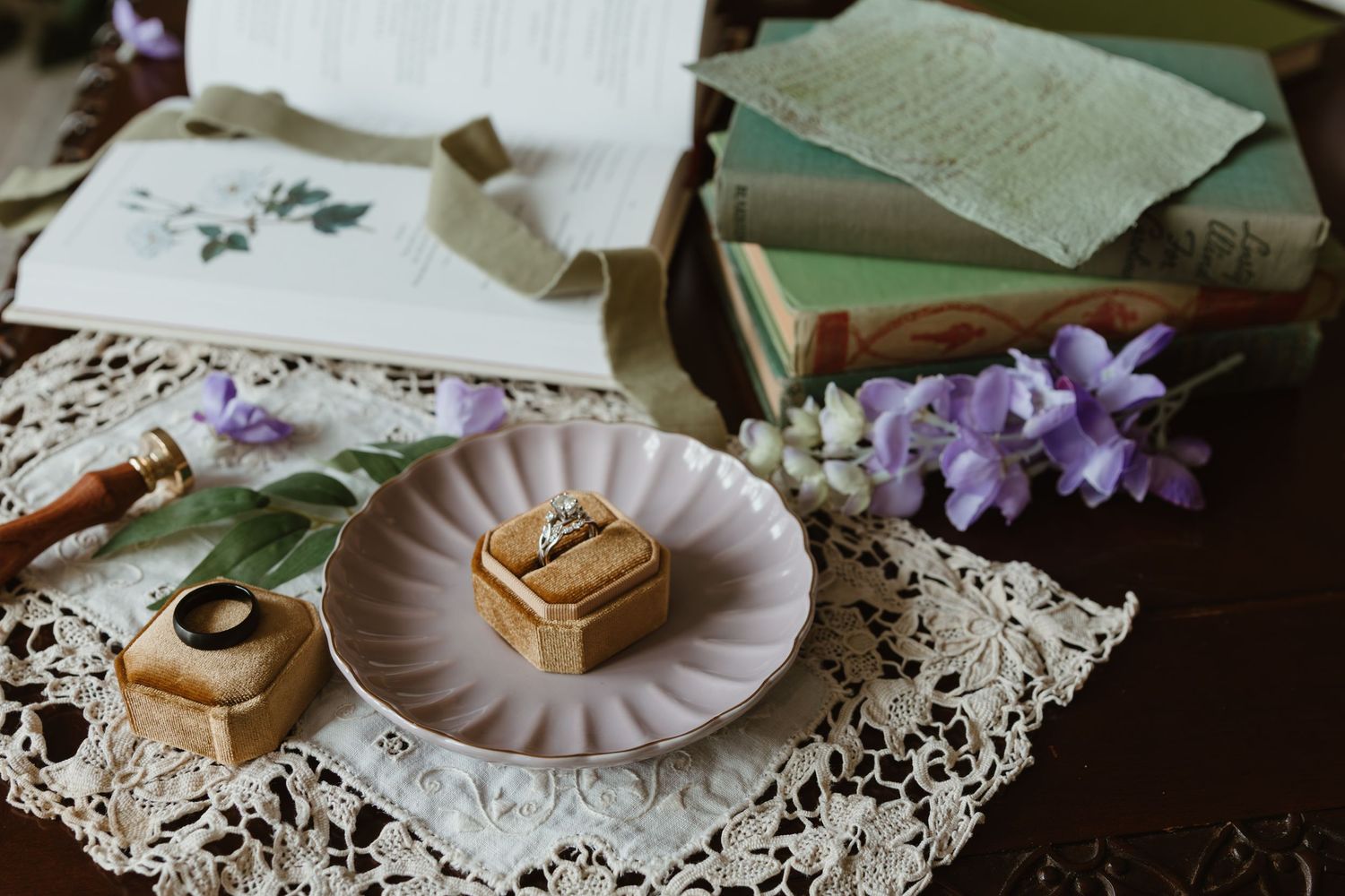 Wedding rings displayed on a decorative plate with antique lace and purple flowers on a wooden table.