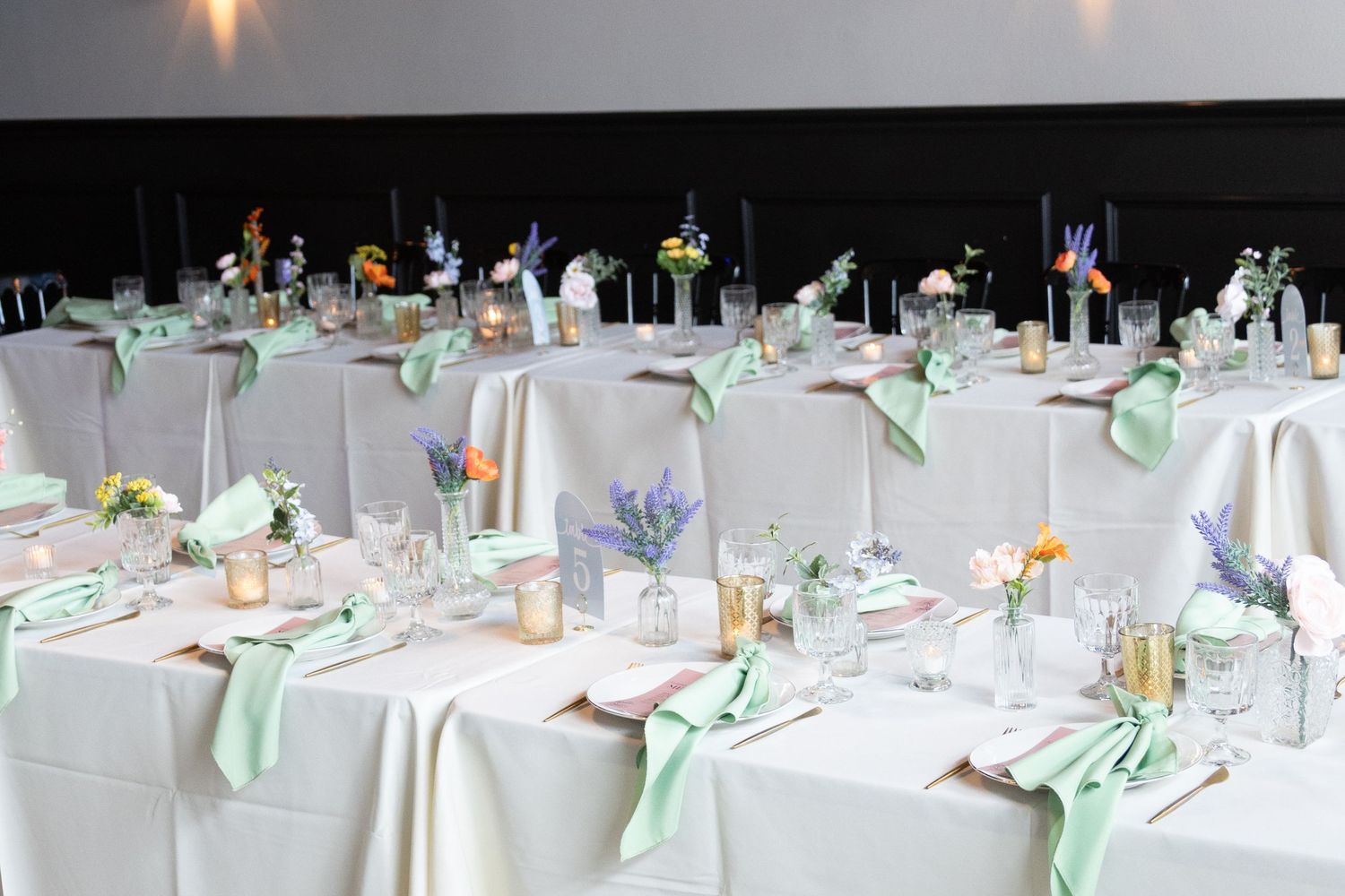 Elegant wedding reception tables with white linens, mint green napkins, and floral centerpieces in a dimly lit banquet hall.