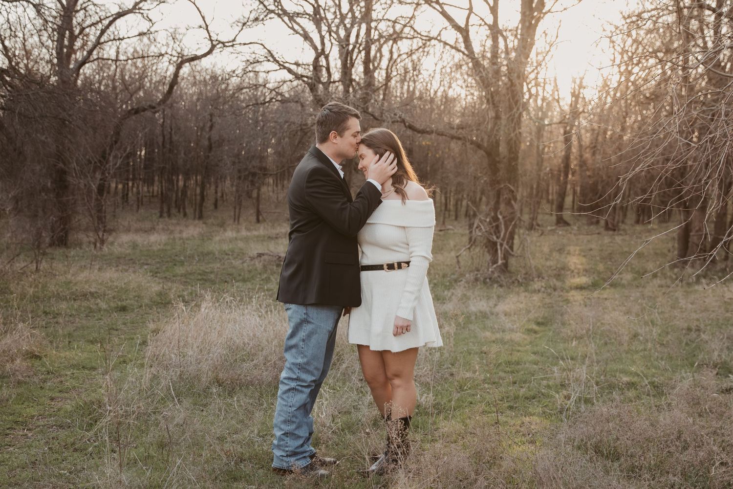 Couple sharing a romantic moment at golden hour in a rustic field surrounded by bare trees, woman in white dress and boots.