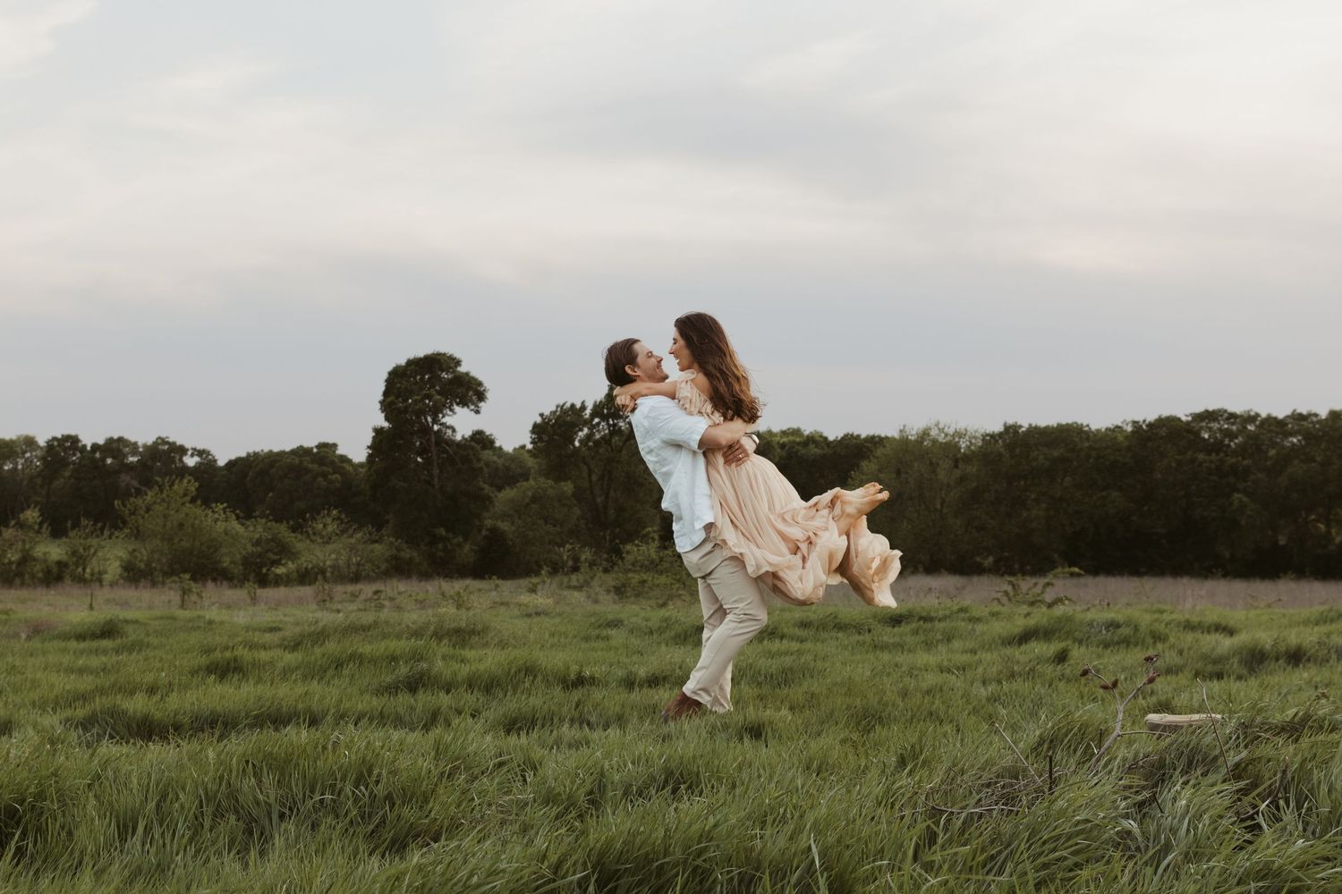 A romantic couple in white attire dances together on a grassy field during sunset.