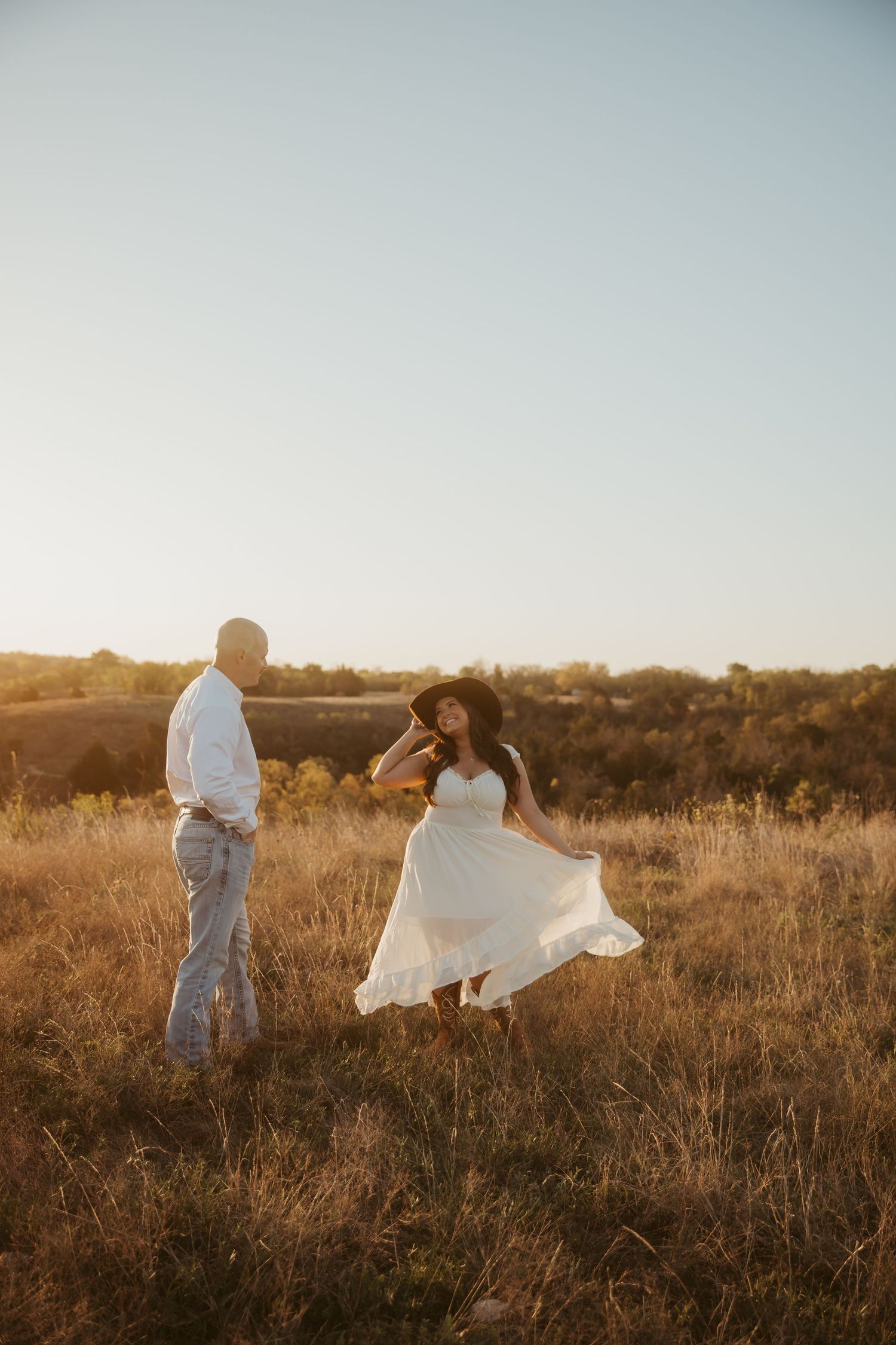 Woman in flowing white dress and wide-brim hat poses in golden field at sunset while man in white shirt watches.