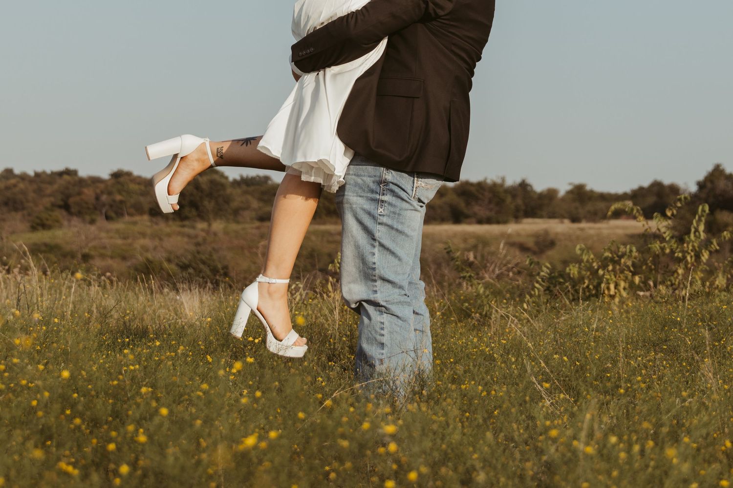 A person wearing jeans and white sneakers stands in a wildflower meadow during golden hour. Engagement photographer DFW