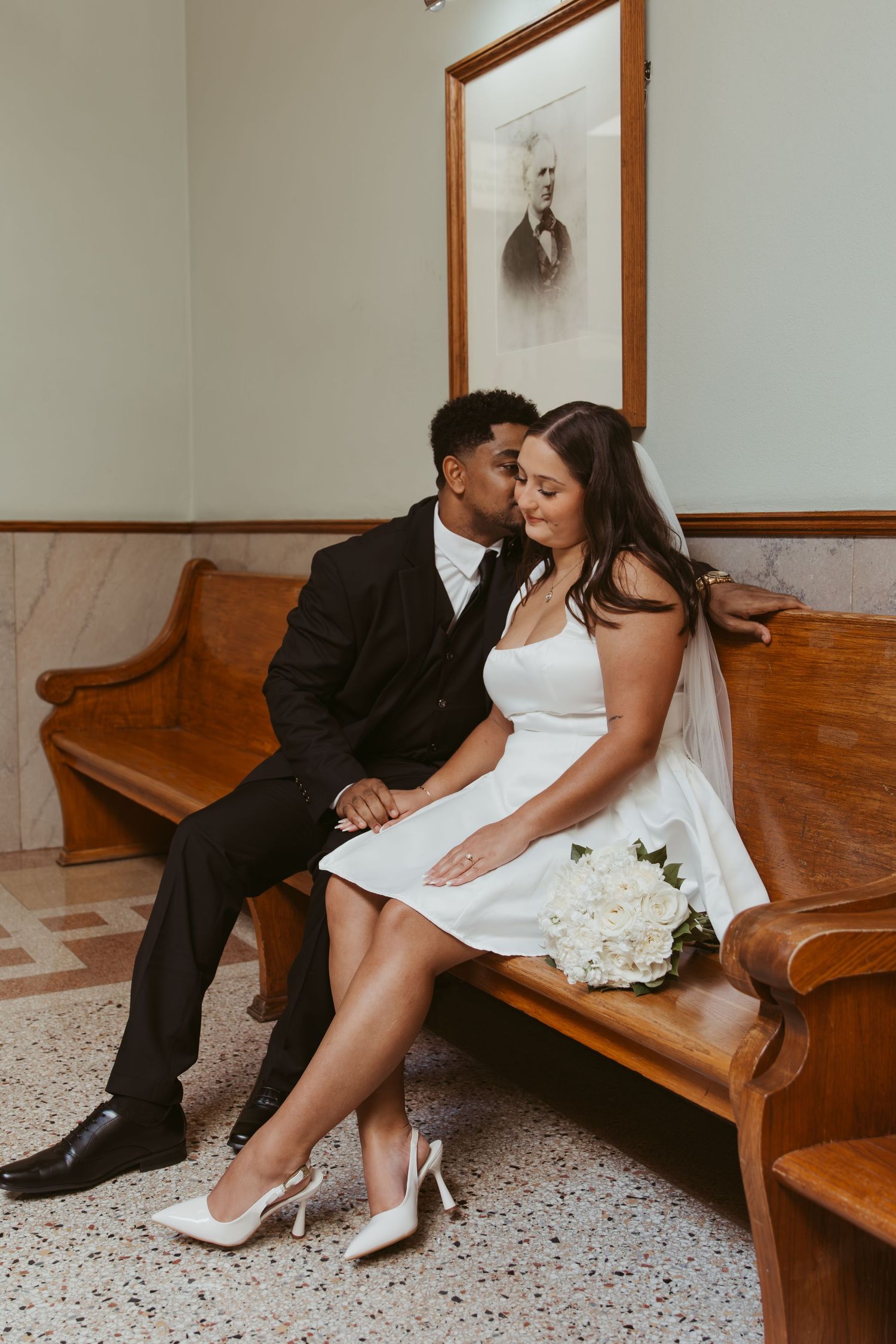 A romantic couple sits close together on a wooden bench in an indoor setting. Wedding Photographer DFW