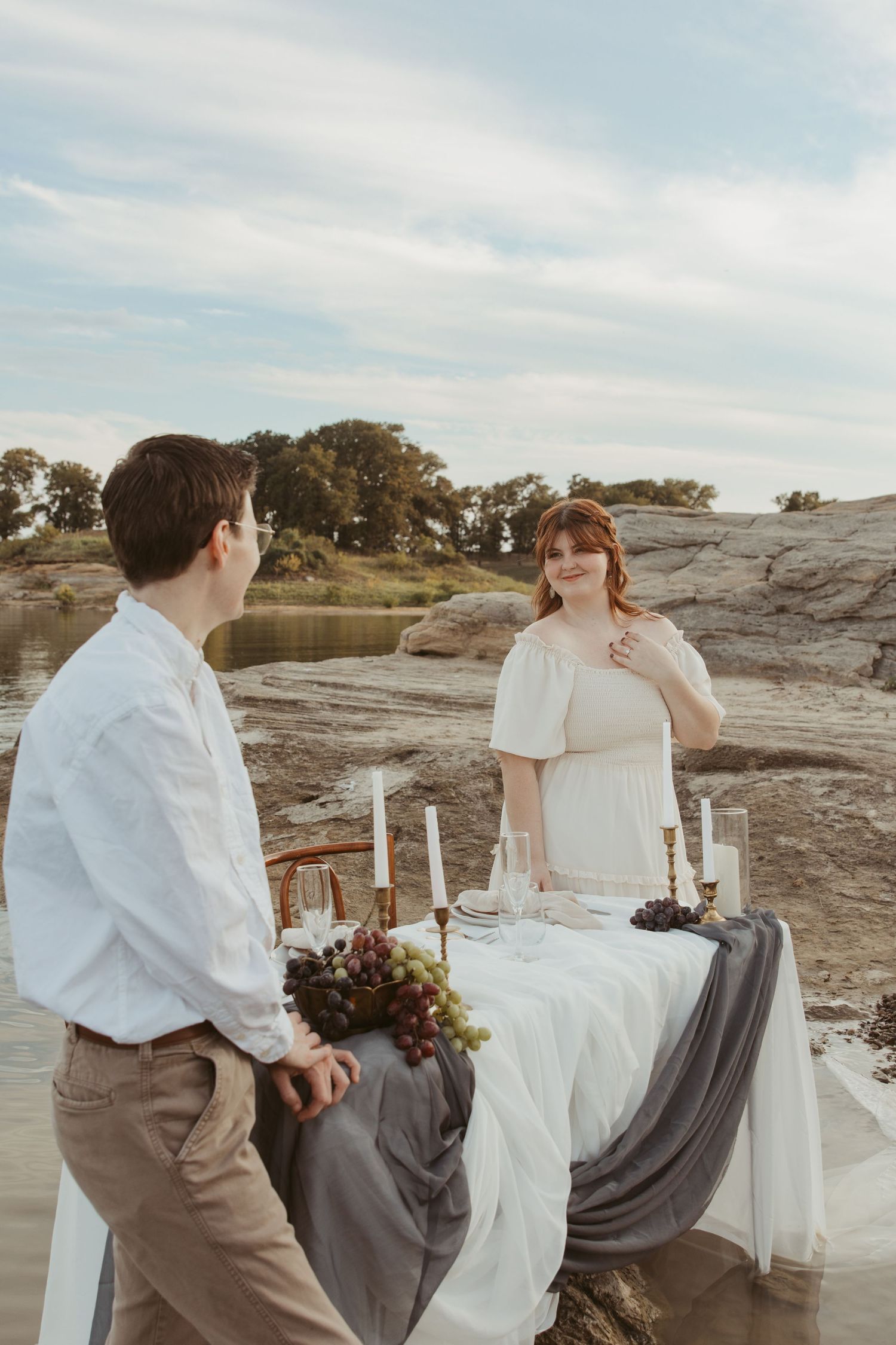 An elegant table setting on rocky terrain by a lake with white candles and rustic decor.