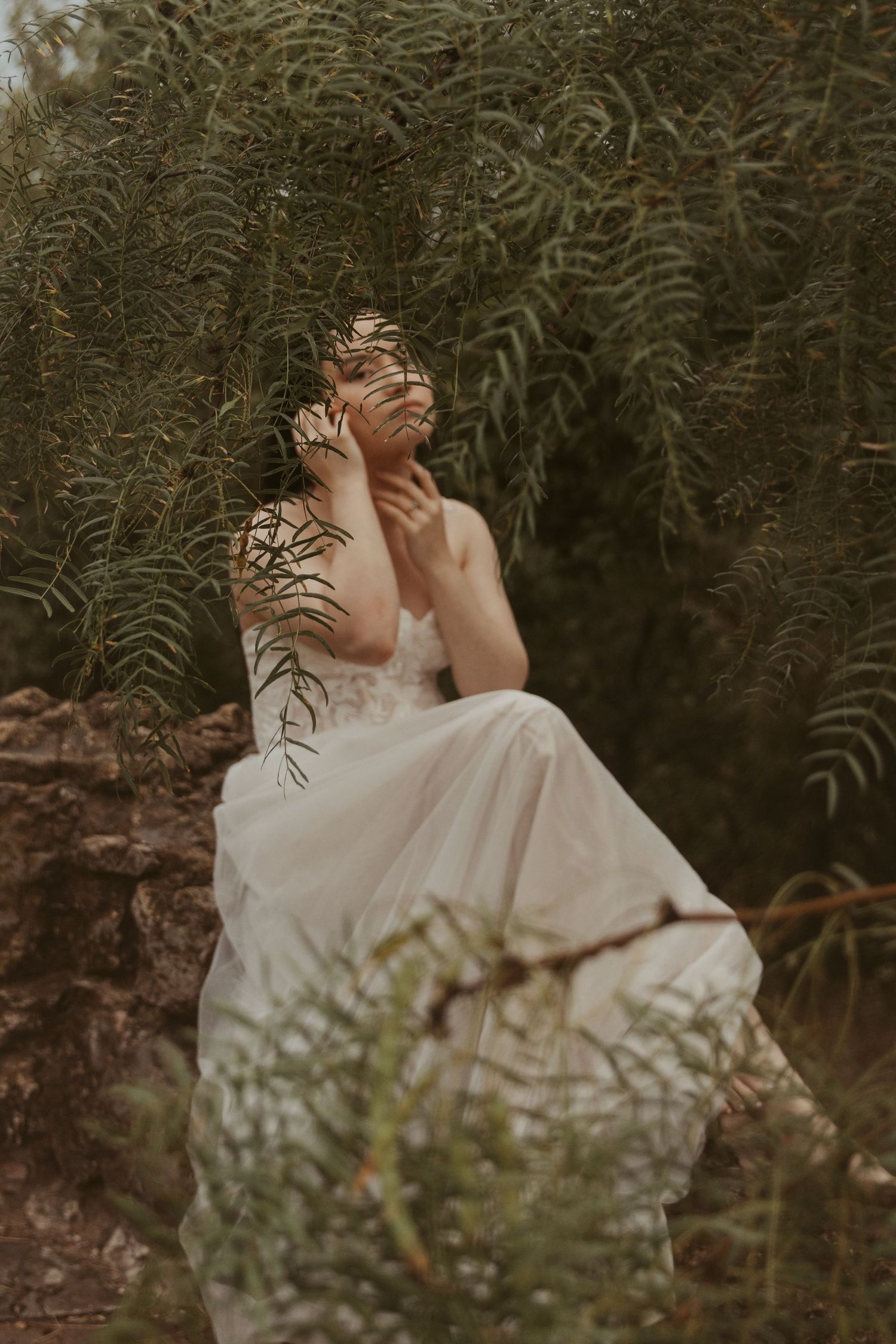 Ethereal portrait of a dreamy figure in a white dress sitting among lush ferns in a moody forest setting.