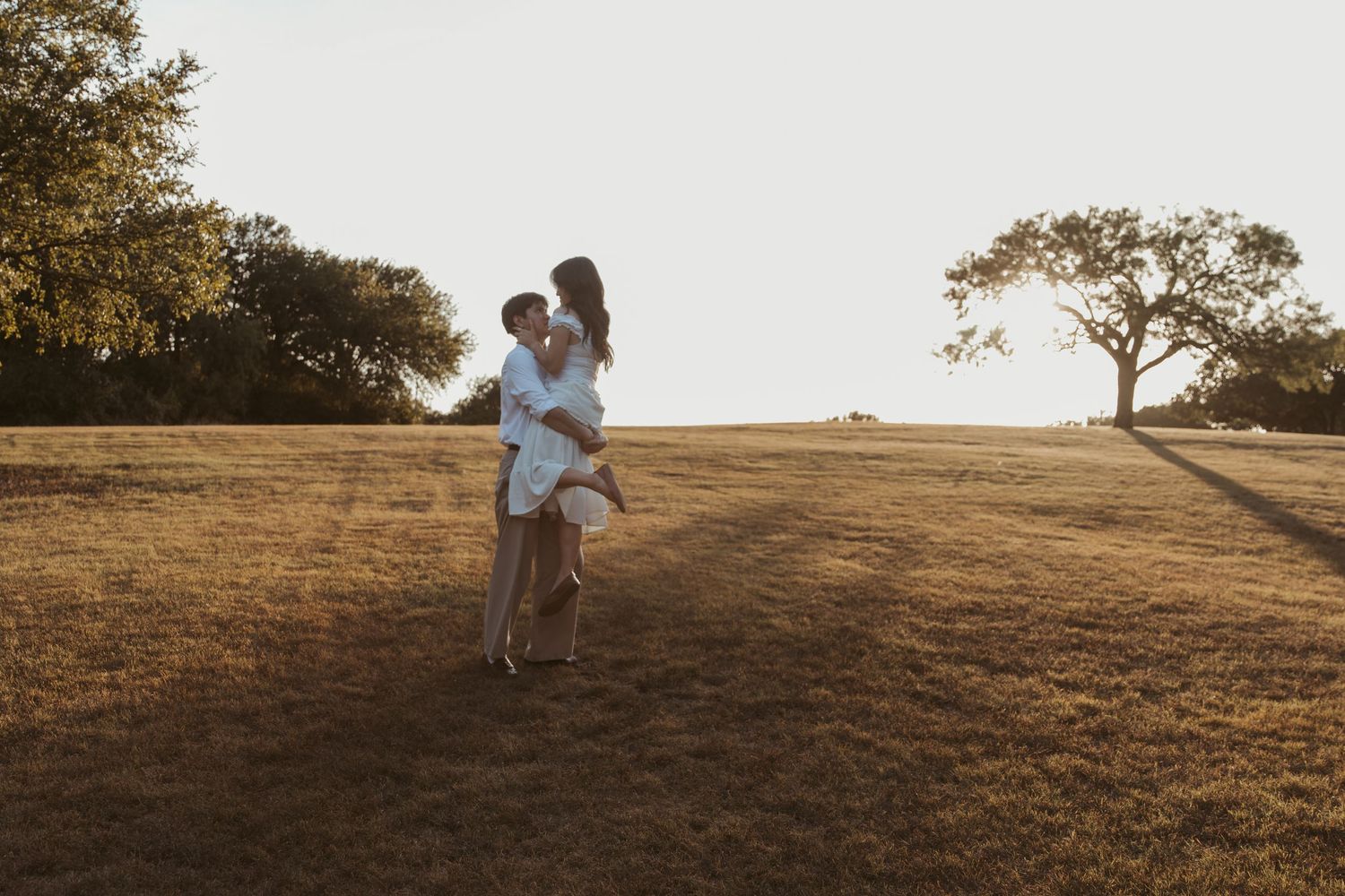 Two figures embrace on a golden hilltop at sunset with trees silhouetted against the warm sky.
