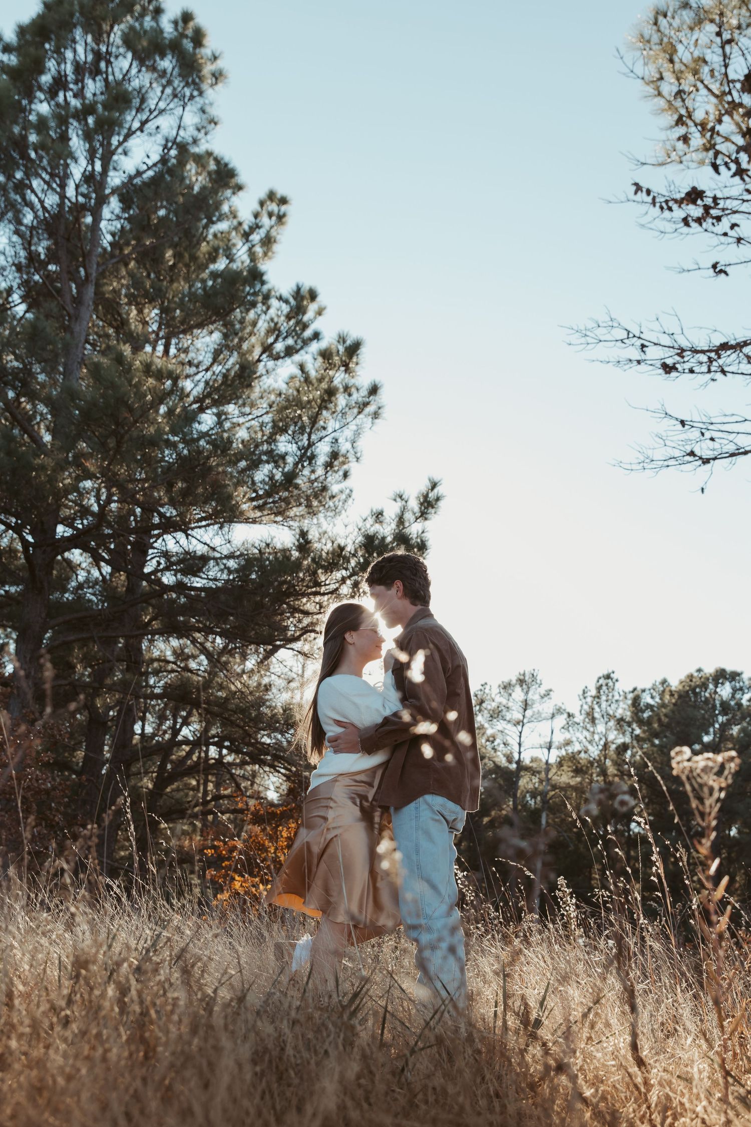 A romantic couple shares tender moments in a golden field with tall grass and pine trees at sunset. engagement photos DFW