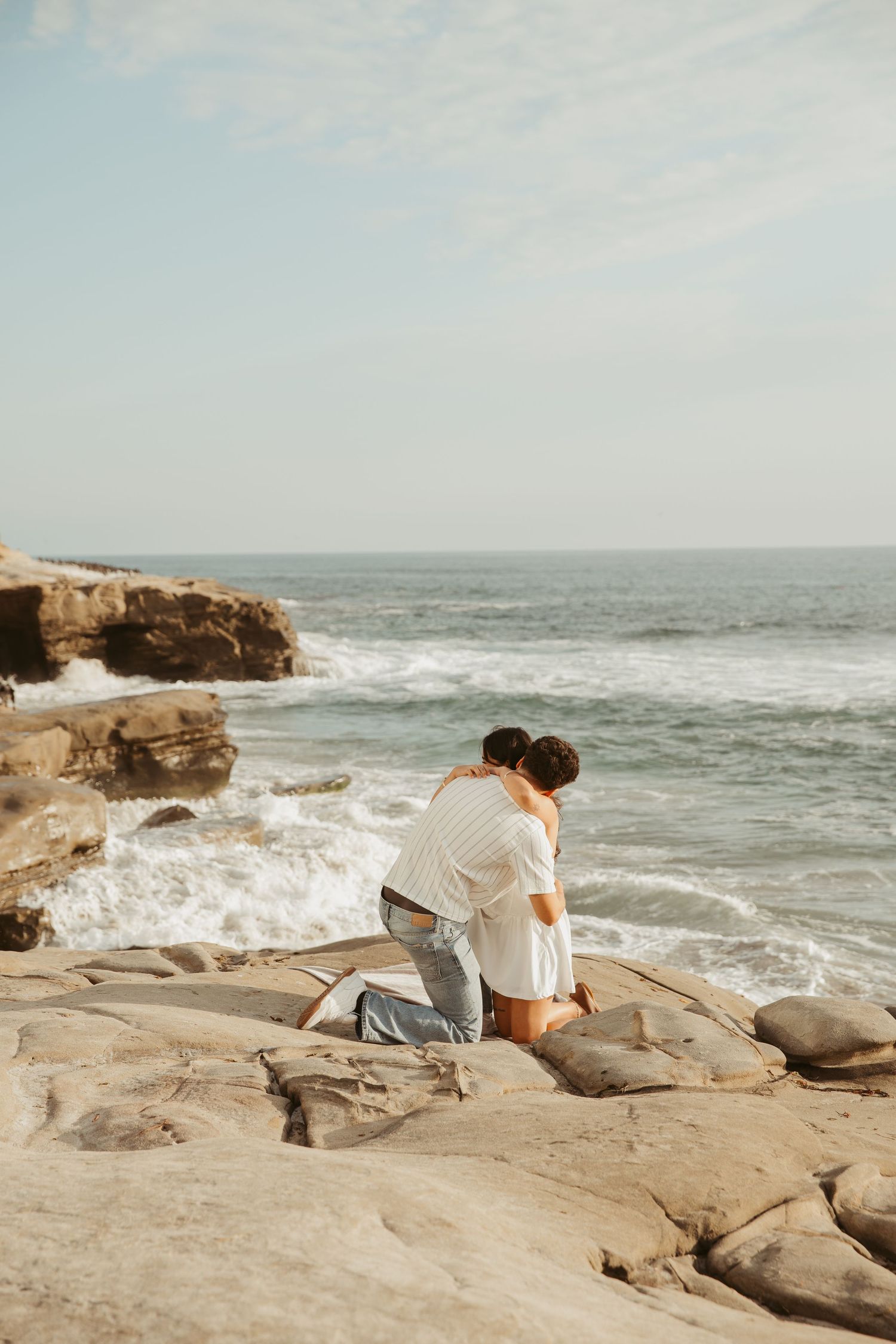 Couple embracing on rocky coastal cliffs with ocean waves crashing behind them on a sunny day.