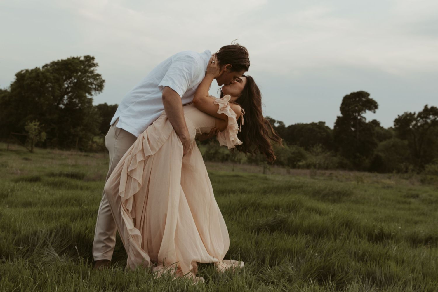 A romantic couple embraces in a dramatic dip kiss against a dreamy pastoral backdrop at sunset.