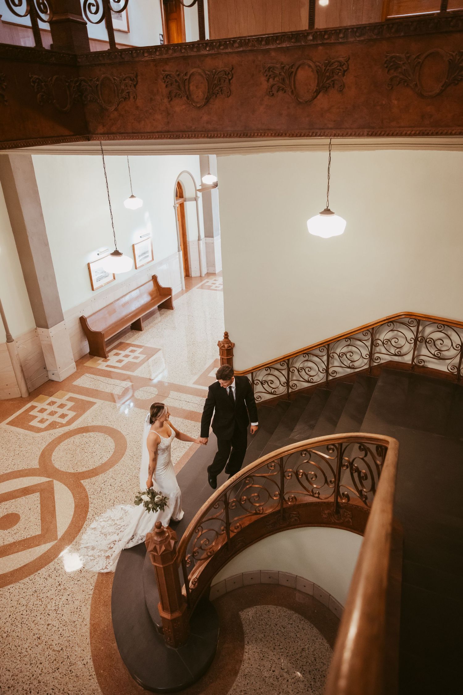 A couple stands on an elegant curved staircase with ornate railings and geometric floor patterns in a vintage building interior.