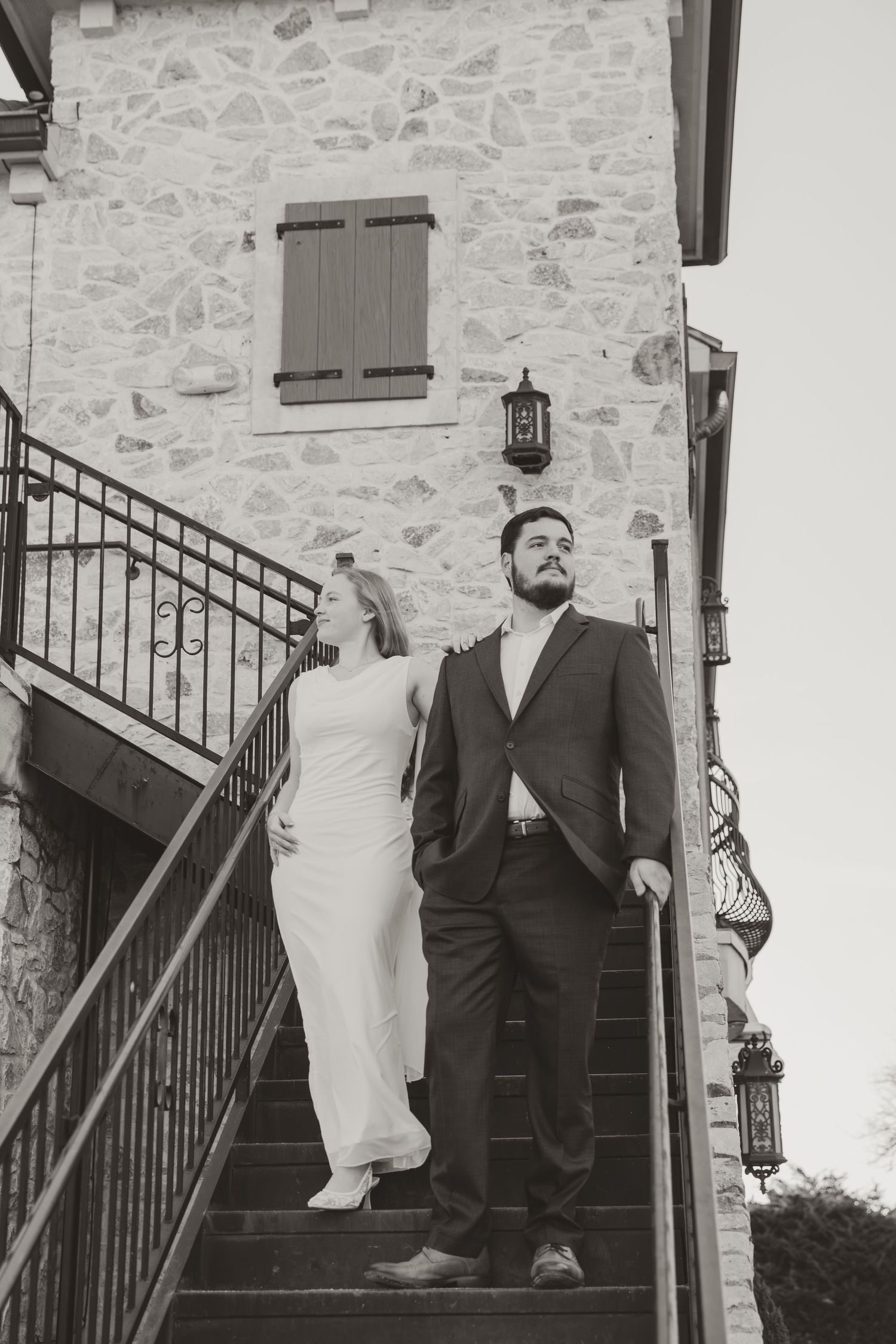 A couple in wedding attire poses on stone stairs against a rustic stone wall with wooden shutters.