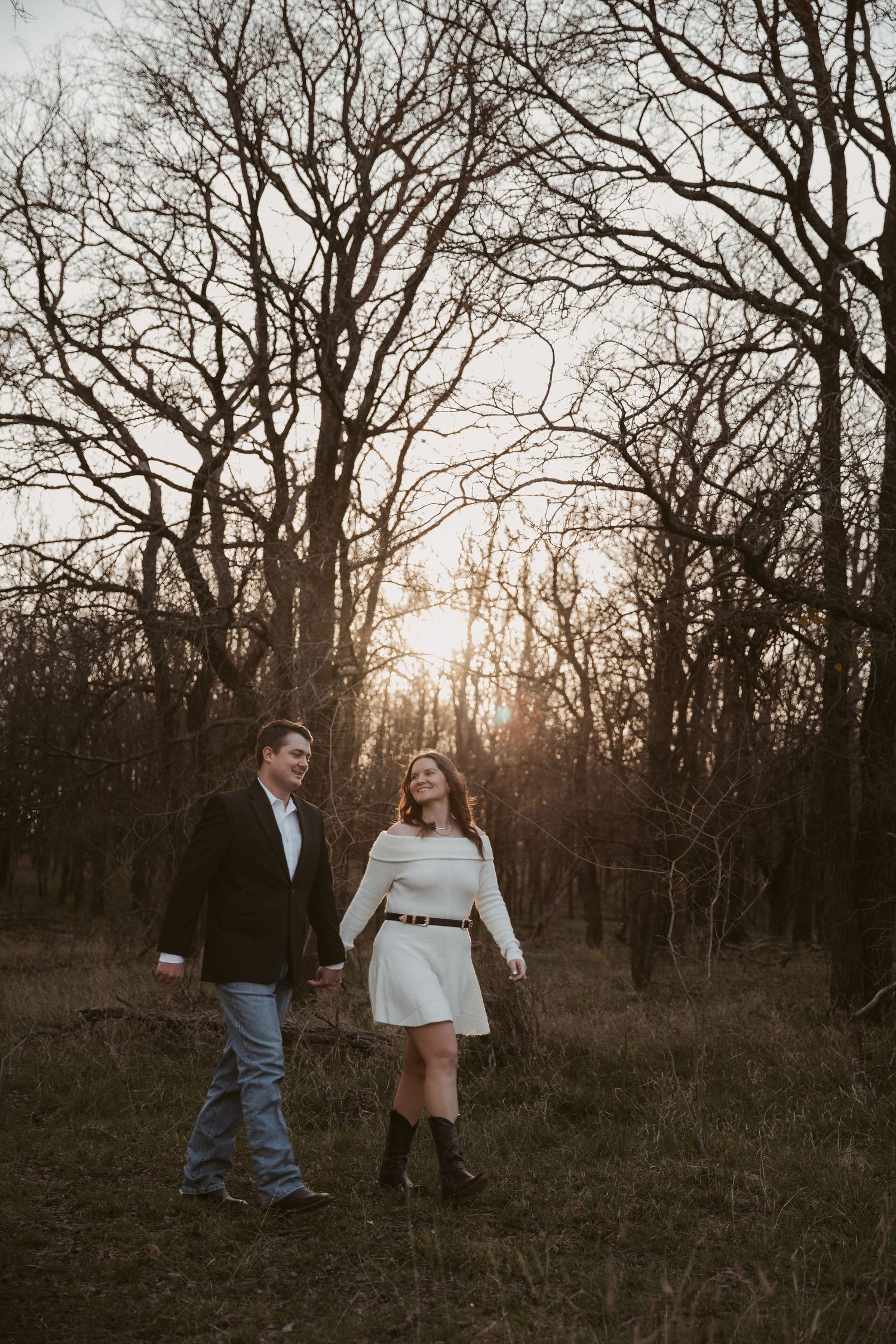 Couple holding hands walking through a golden sunset forest in winter, woman in white dress and cowboy boots.