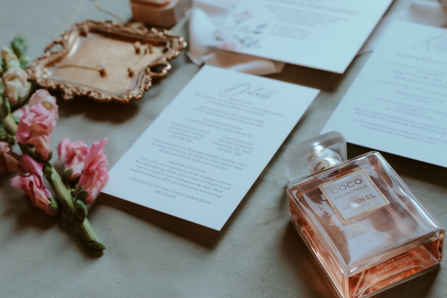 A wedding invitation suite with pink flowers and perfume bottle arranged on a grey surface.