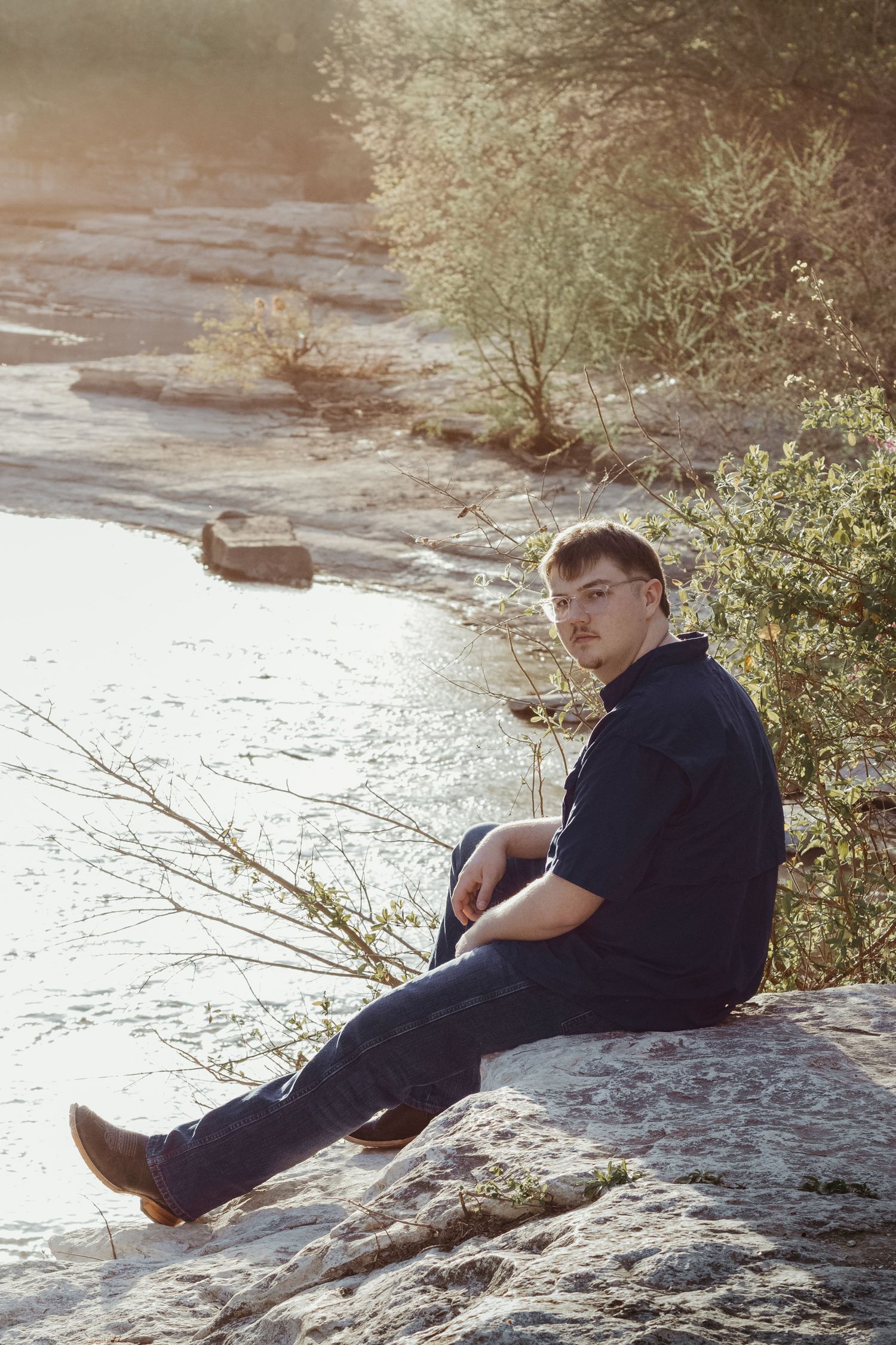 Person sits on rocky riverbank during golden hour sunlight with trees and flowing water in background.