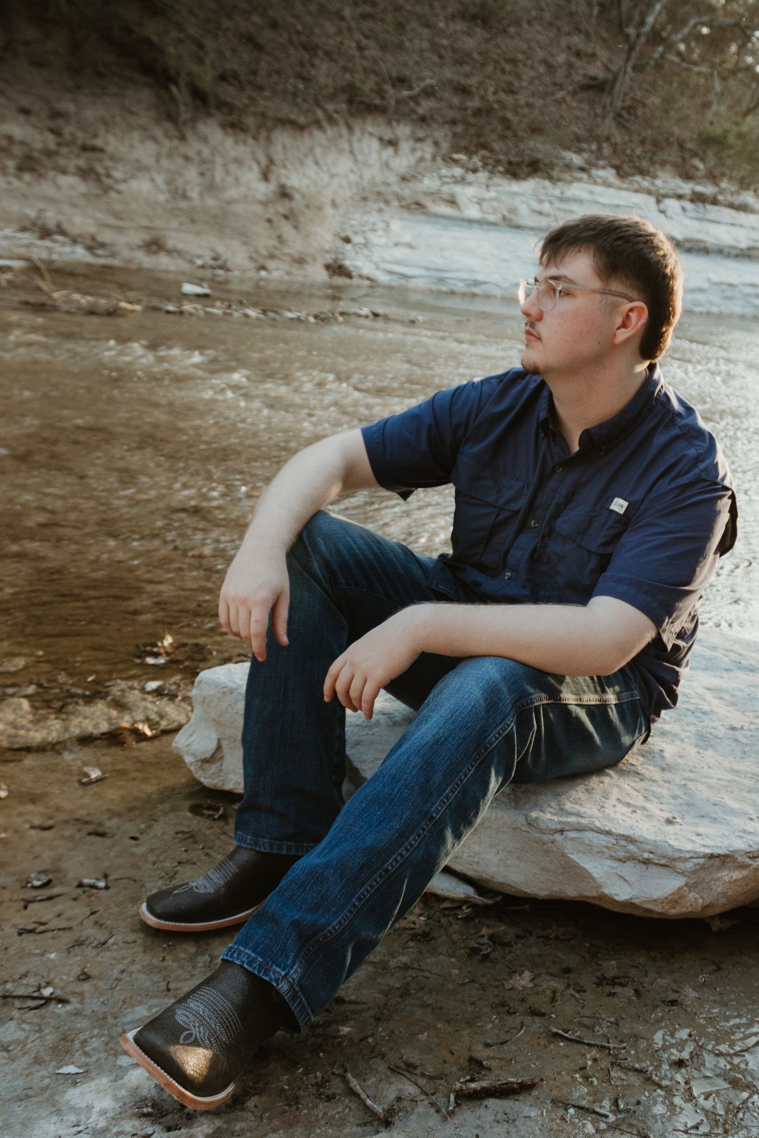 Person sits on a rock ledge wearing a navy button-down shirt and jeans in a natural outdoor setting at dusk.