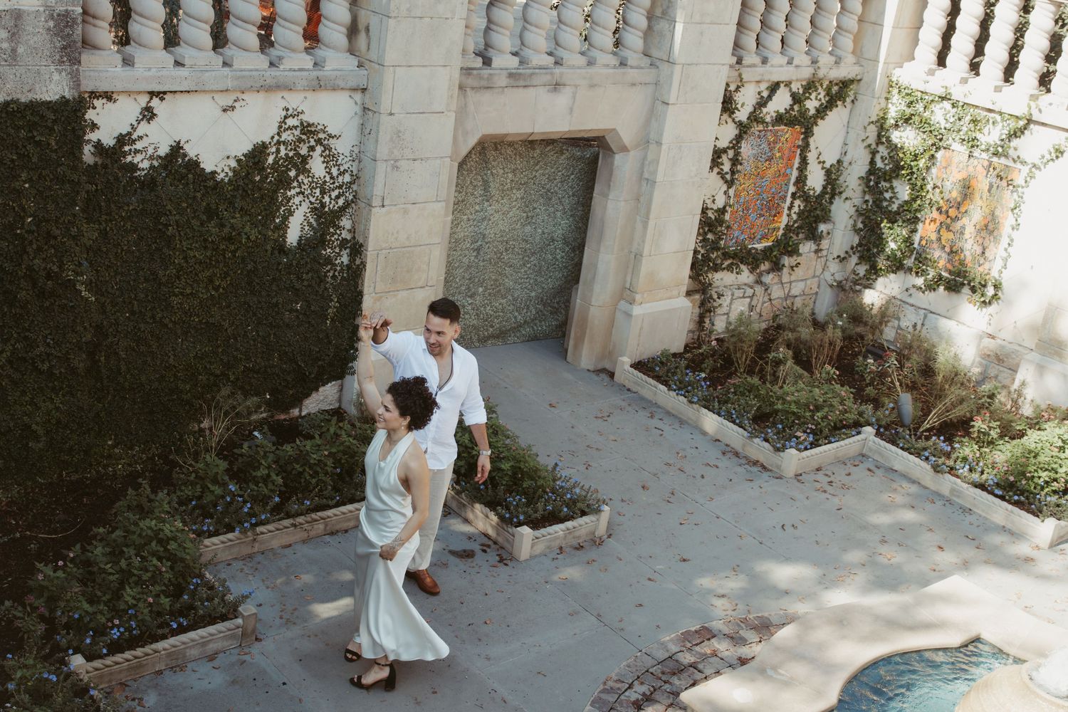 A person in a white dress walks through a stone archway in a Mediterranean-style courtyard setting.