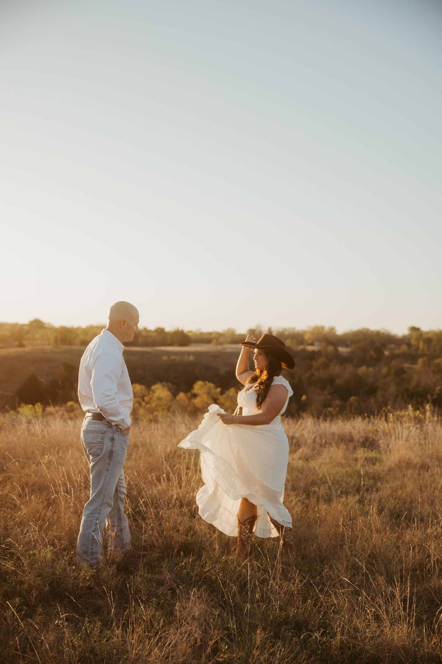 Couple dancing in a sunlit field at golden hour, woman in flowing white dress holding a cowboy hat.