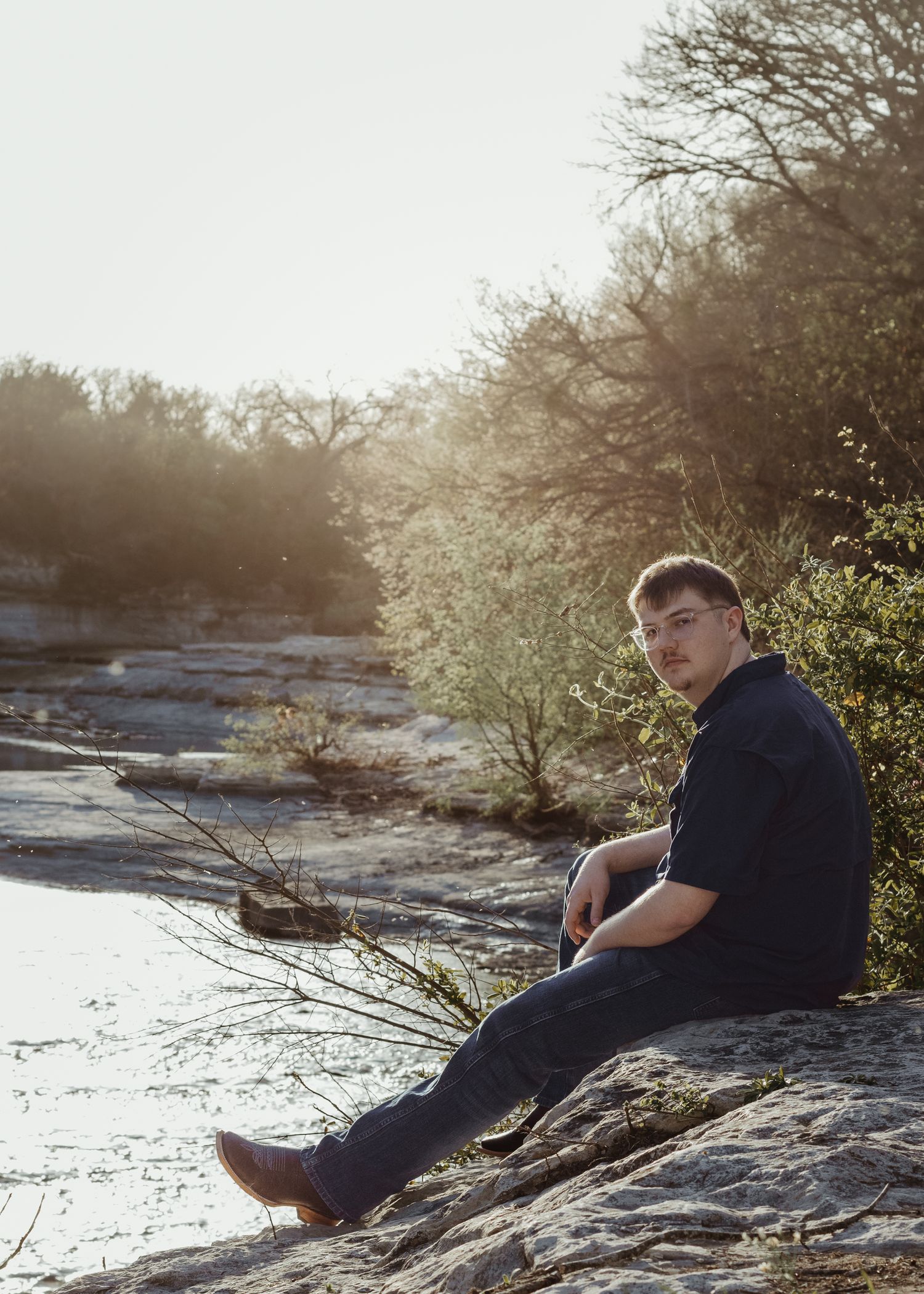 Someone sits on a rocky riverside during sunset as golden light filters through bare winter trees and reflects off the water.
