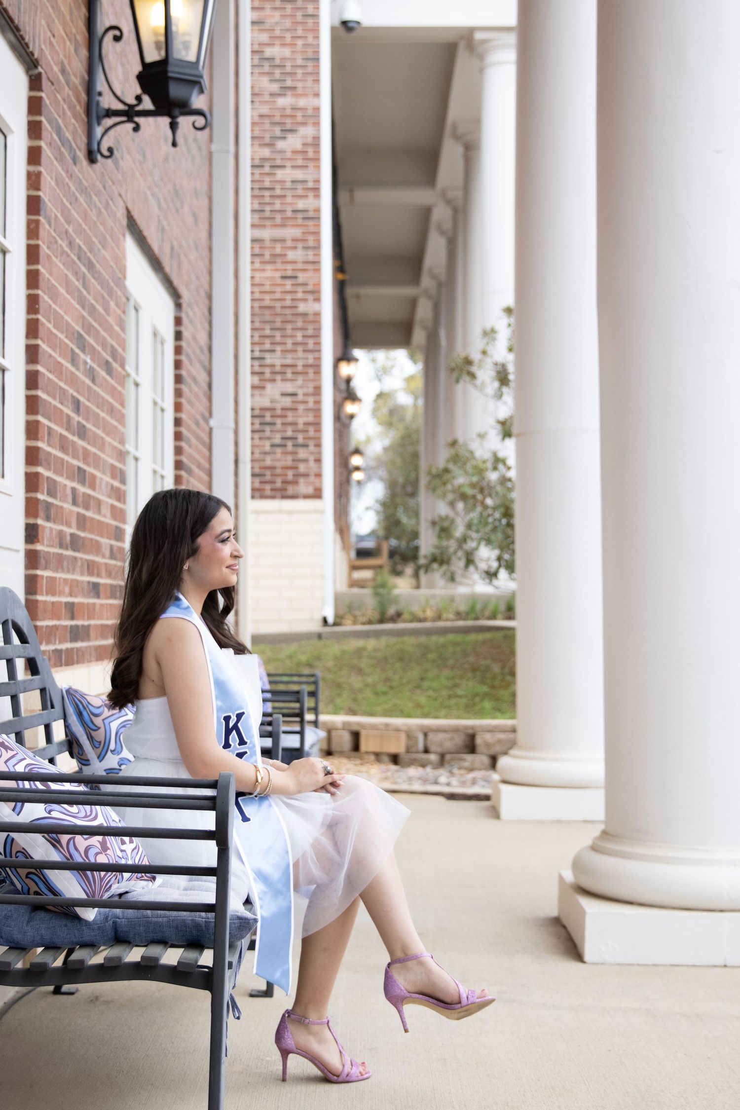 A person in a white dress sits on a bench outside a brick building with stately white columns and lanterns.