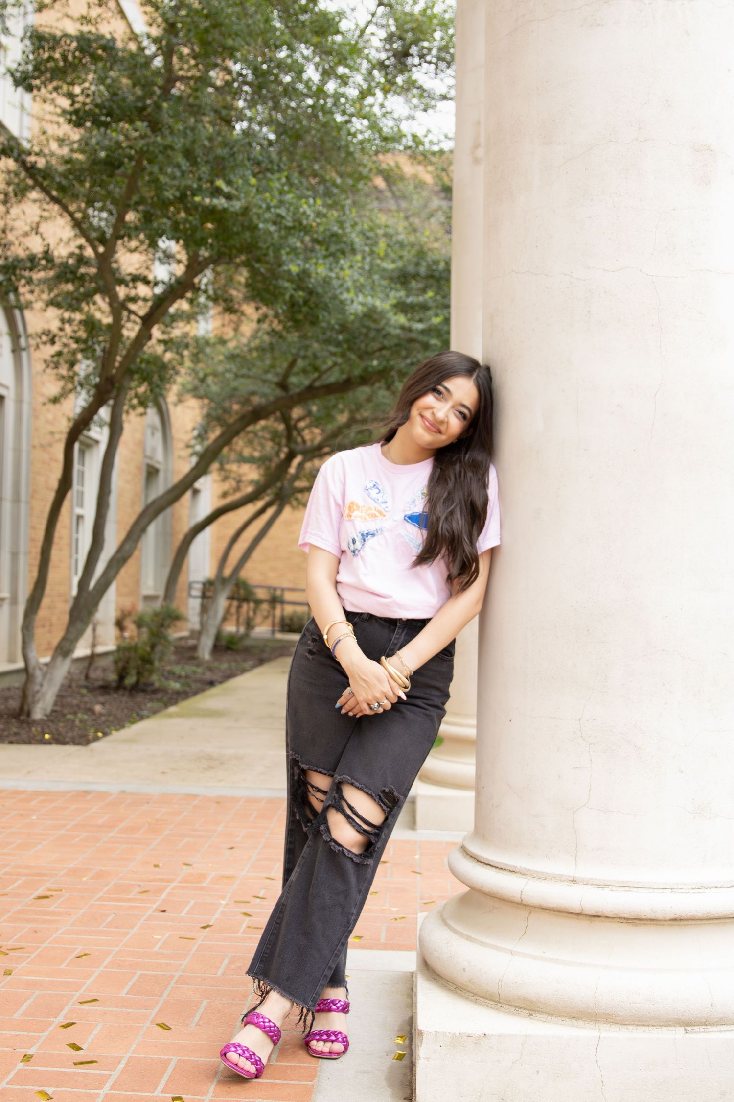 Woman in casual attire with ripped black jeans and pink sandals poses against white column outdoors on brick pathway.