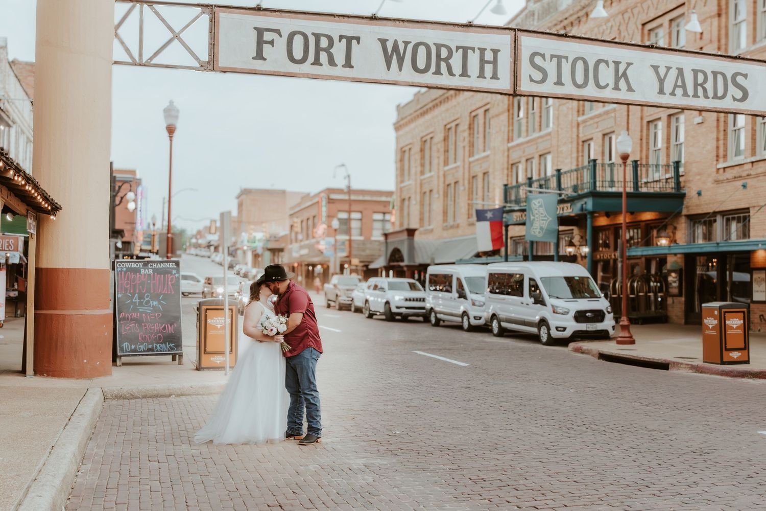 A couple embraces on a brick sidewalk outside the historic Fort Worth Stock Yards in Texas with vintage buildings in view.