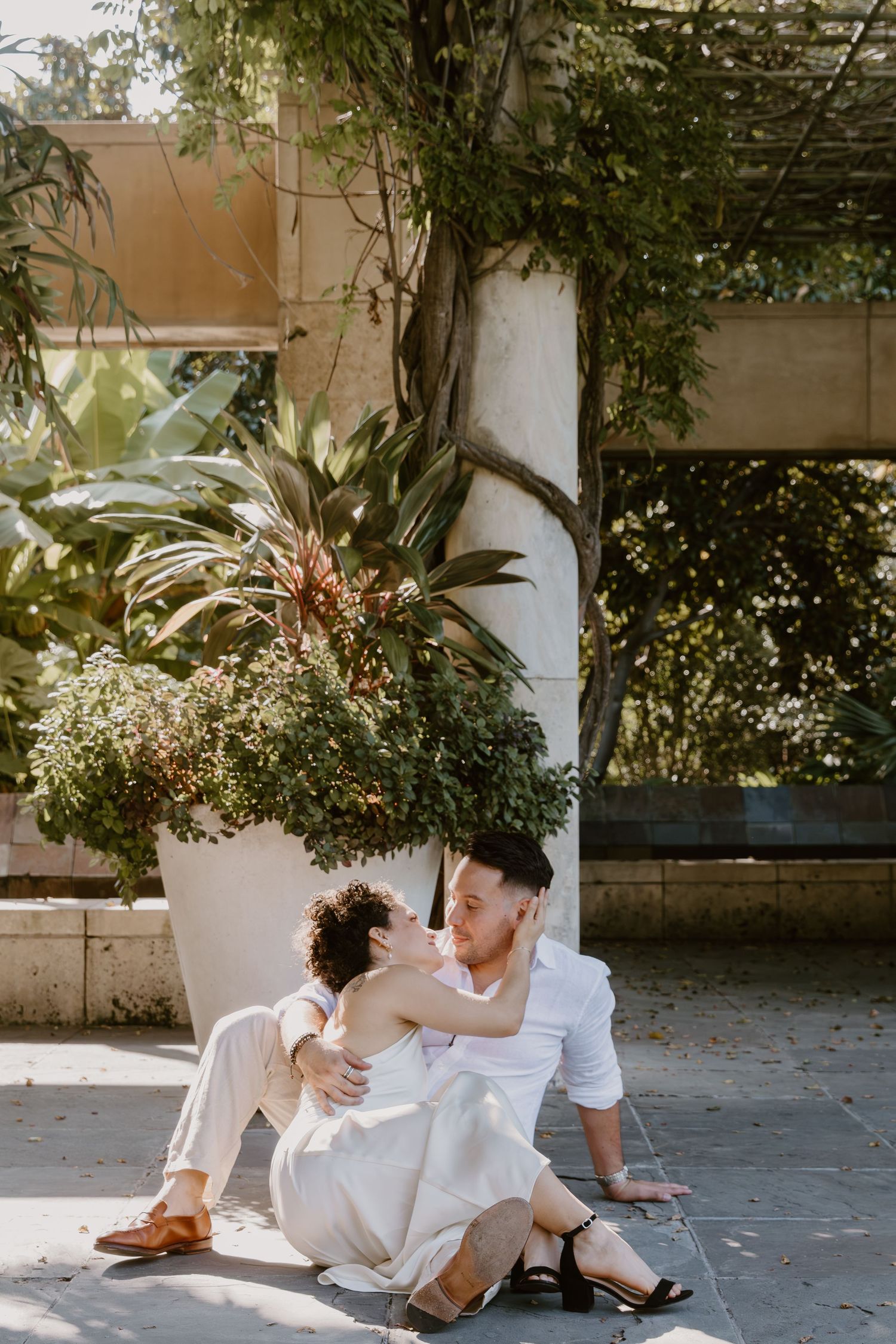 A romantic couple in white outfits lounges together on a paved outdoor surface surrounded by greenery.