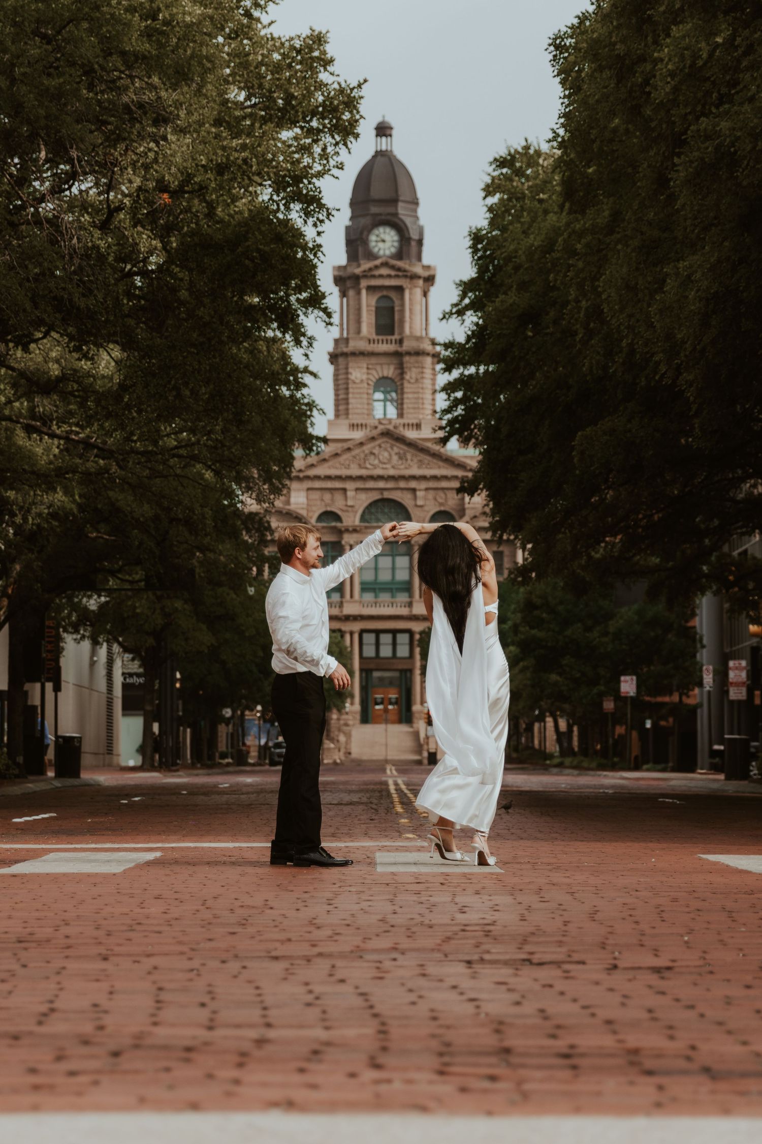 A couple poses together on a brick pathway with a grand historic courthouse clock tower in the background.