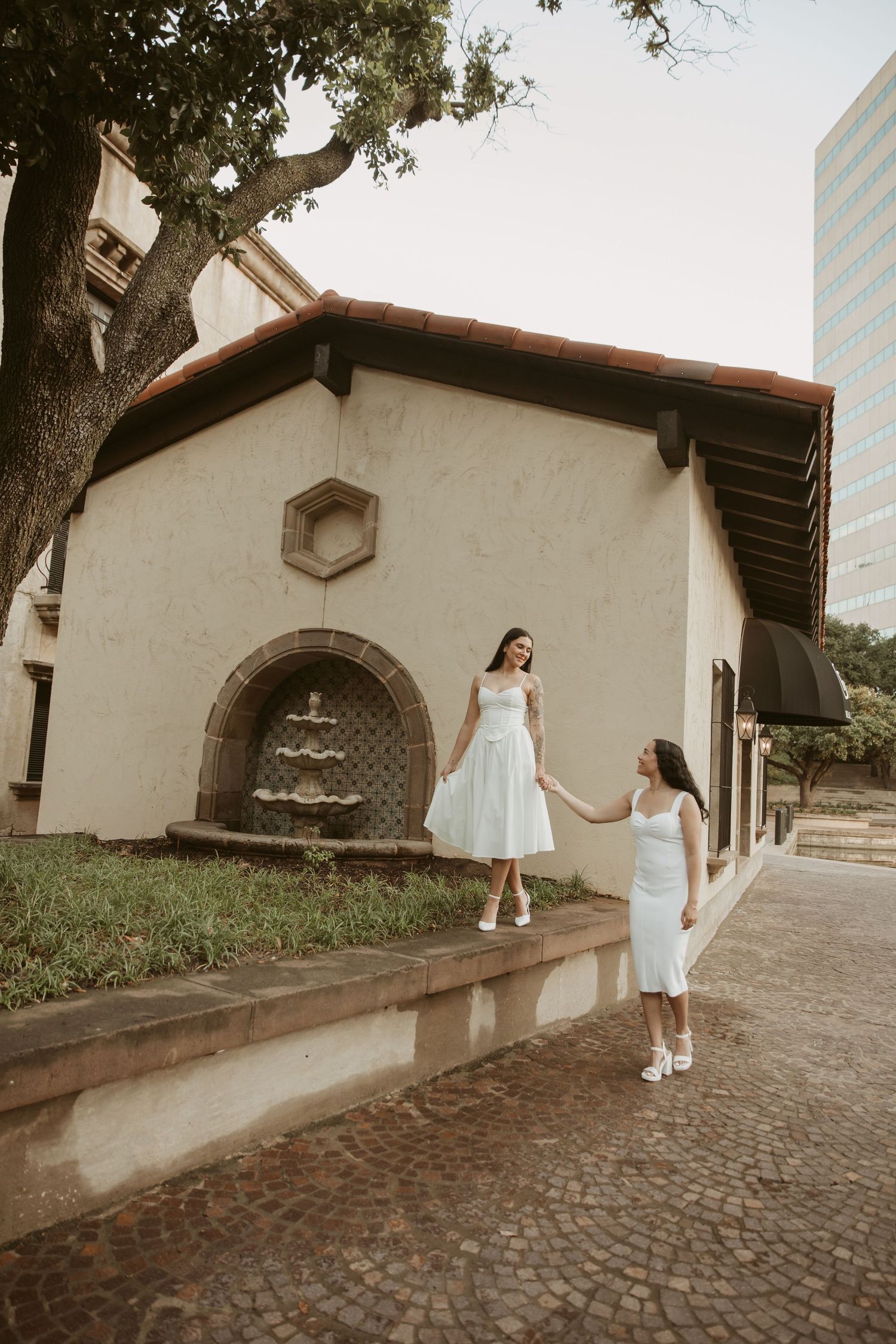 Two women in white dresses hold hands outside a historic Spanish-style building with a fountain on a cobblestone street.