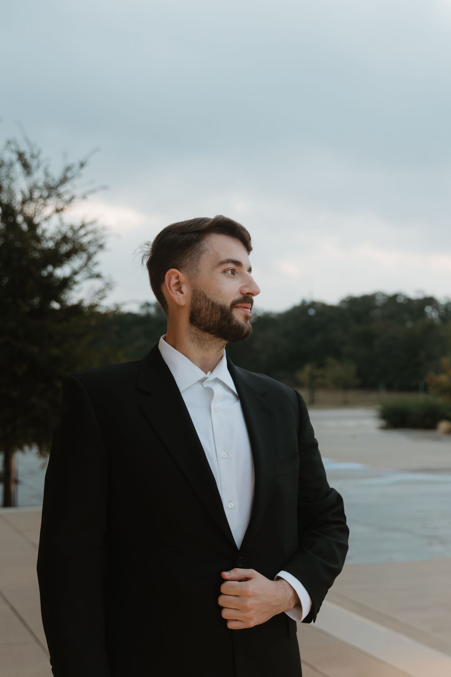 A groom in a black suit and white tie poses outdoors against a waterfront backdrop during sunset. Wedding Photographer Fort Worth