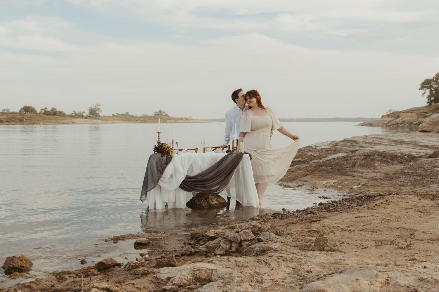 Couple in wedding attire poses together along a scenic waterfront beach at sunset with white tent in background.