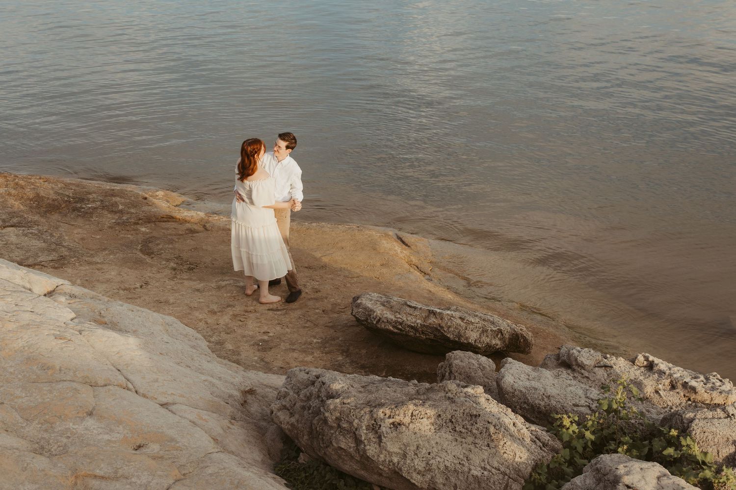 A couple shares a romantic moment on a rocky beach shoreline at sunset.
