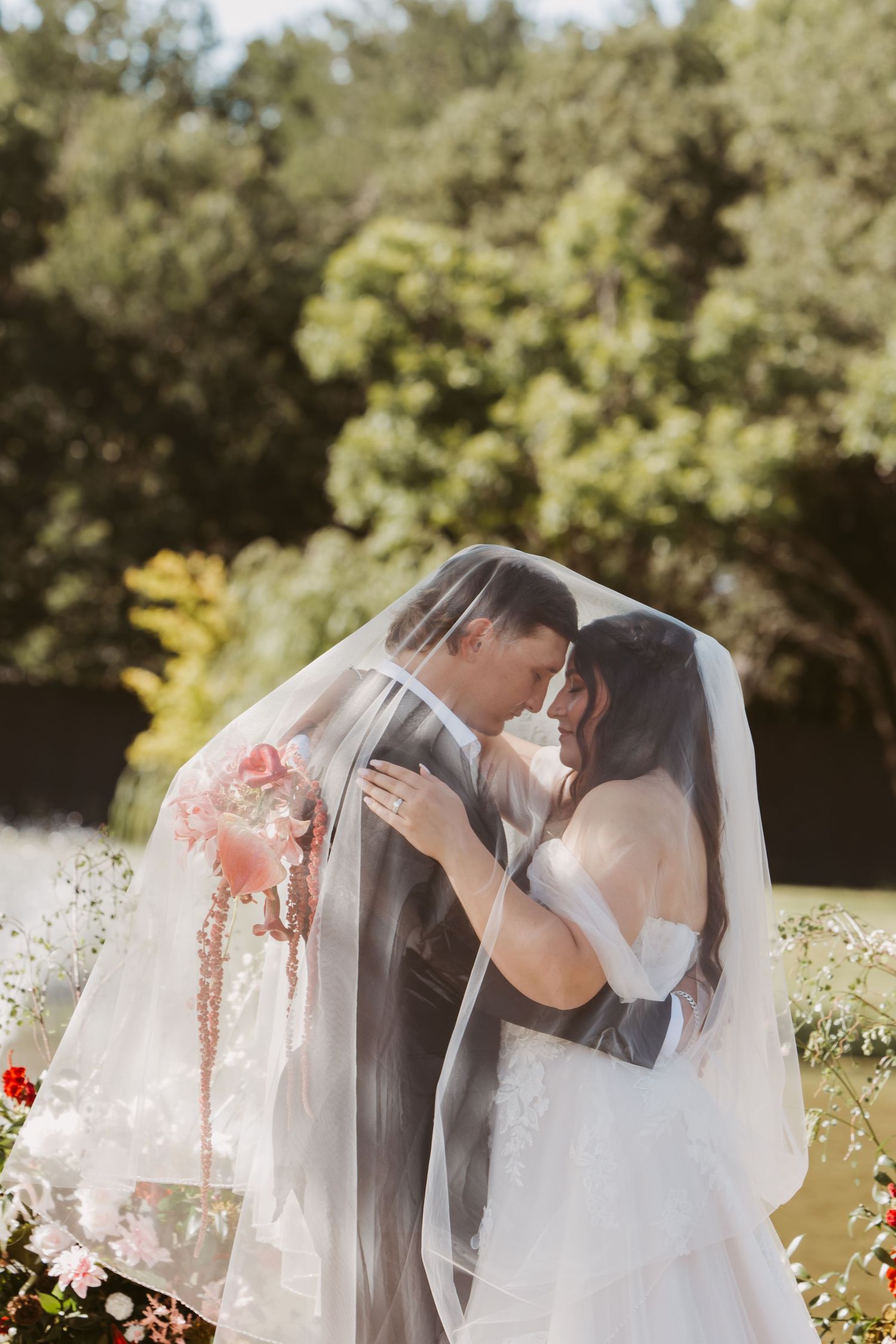 Bride and groom share a romantic moment under a flowing veil outdoors, surrounded by lush greenery and pink florals.