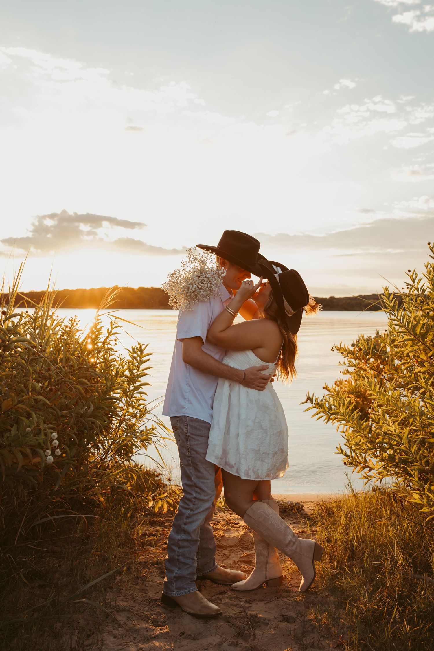 Couple kissing at golden sunset by a lake, woman in white dress and cowboy hat, man holding wildflowers.