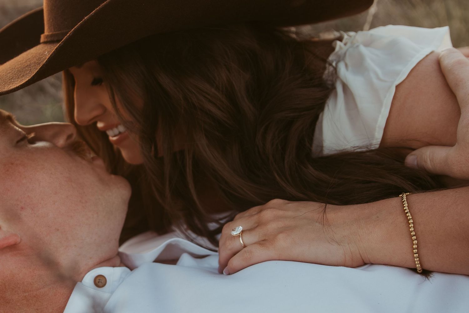 Couple sharing a romantic moment, woman wearing engagement ring and gold bracelet, man in cowboy hat, outdoor photo.