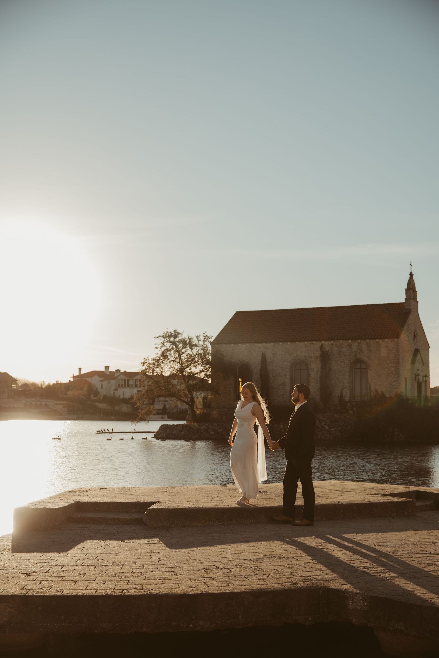 A couple walks along a stone pathway by the sea with a small church and coastal landscape in the background.