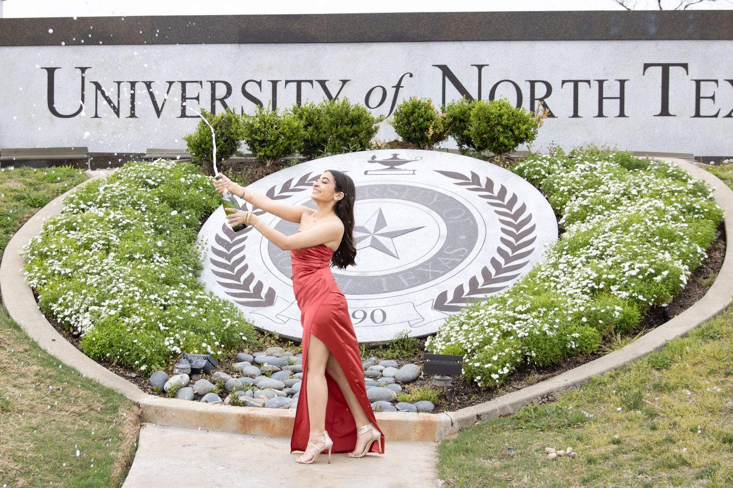 A celebratory graduation photo in front of the University of North Texas seal surrounded by white flowers.