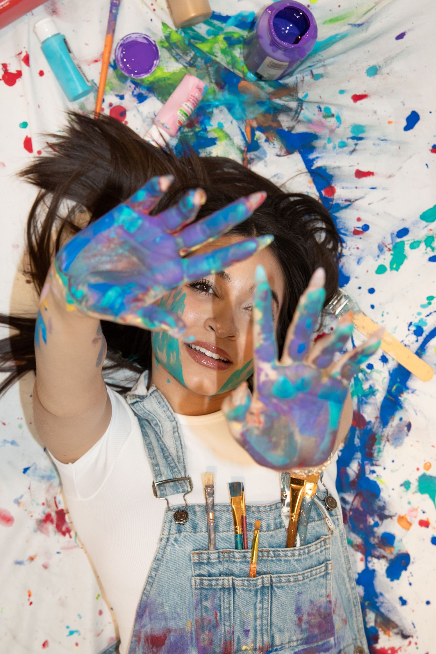 Paint-covered hands playfully cover face against backdrop of vibrant paint splatters and art supplies.
