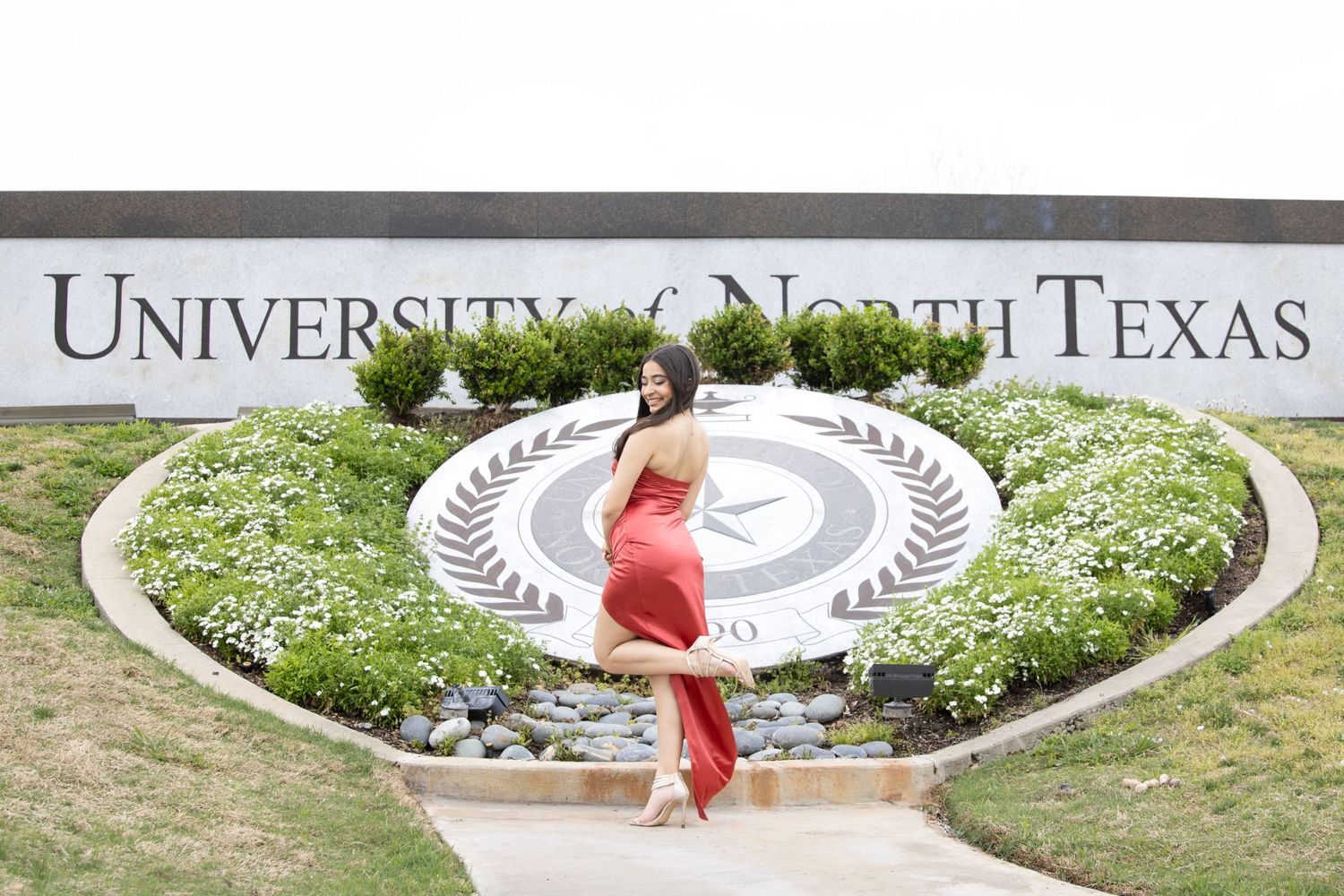 A person in a red dress poses joyfully in front of the University of North Texas entrance sign and landscaping.