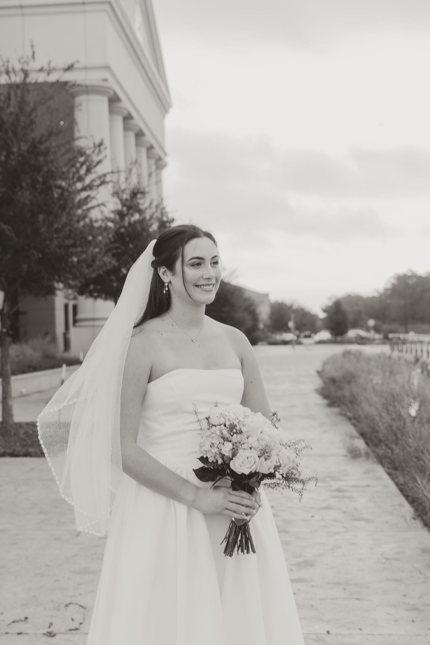 A bride in a flowing white wedding dress holds a bouquet while walking along a stone pathway on an overcast day.