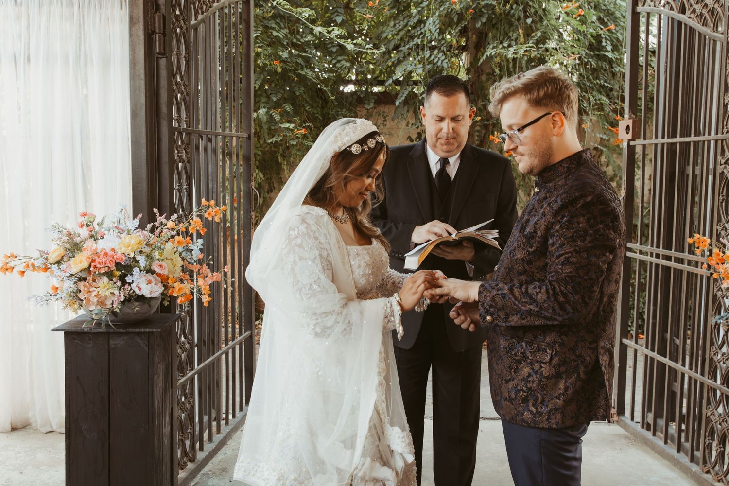 A wedding ceremony takes place in front of a decorative iron gate with autumn foliage in the background.Wedding Photographer DFW