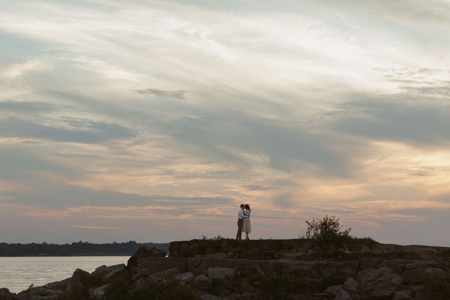 A silhouetted couple stands on a rocky outcrop against a dramatic sunset sky over water.