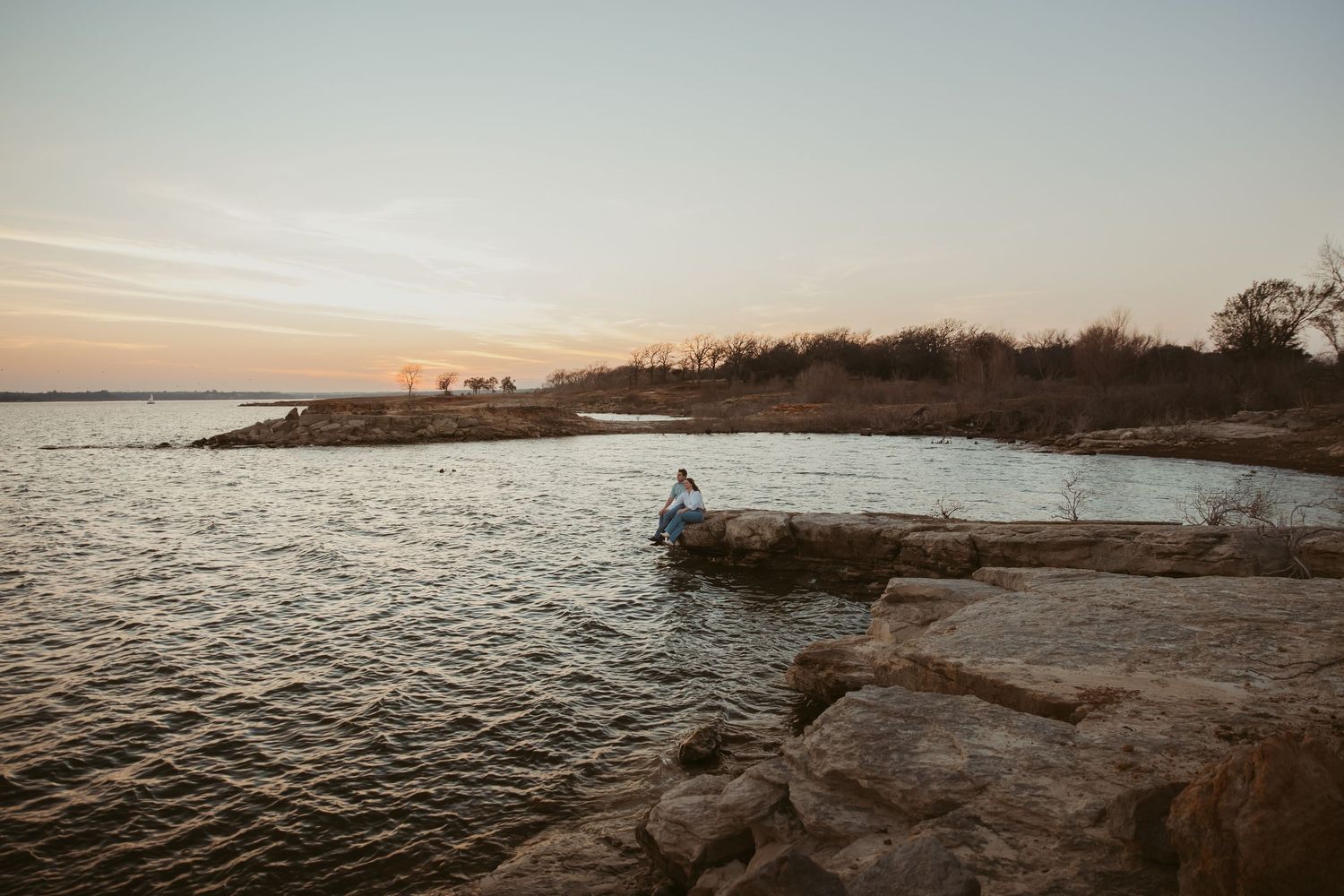 Person sitting alone on rocky shoreline at golden hour sunset beside a tranquil lake in nature.
