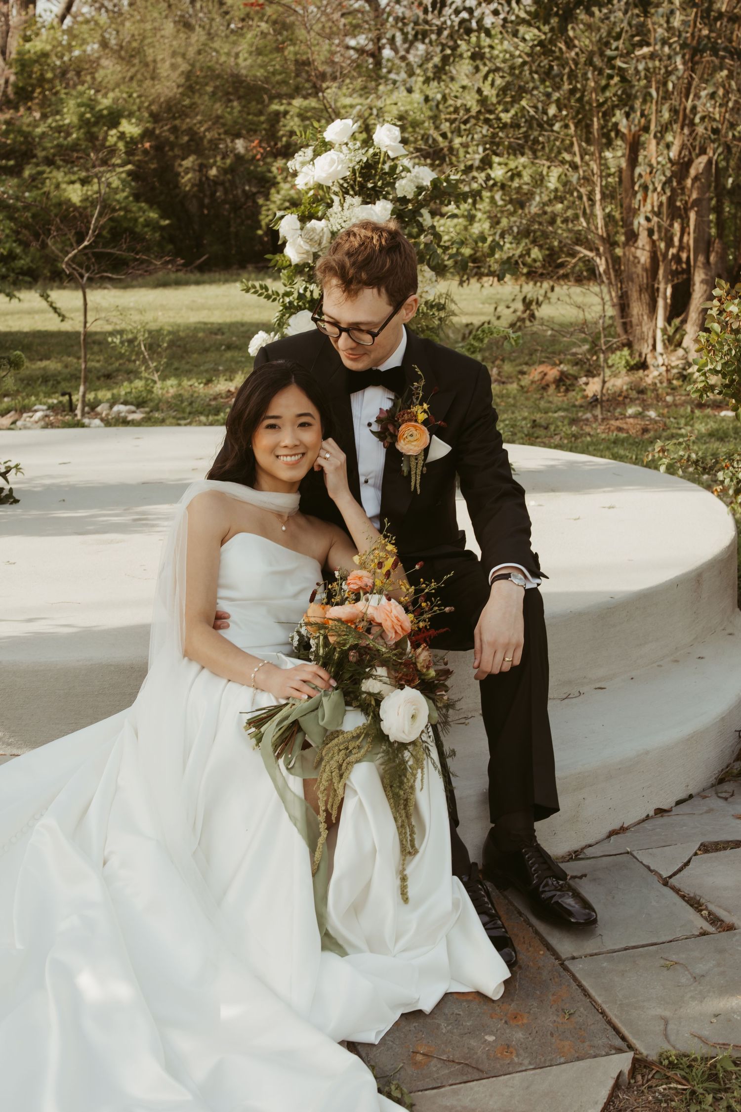Bride in strapless white gown holds bouquet while groom in black tuxedo sits beside her in an outdoor garden setting.